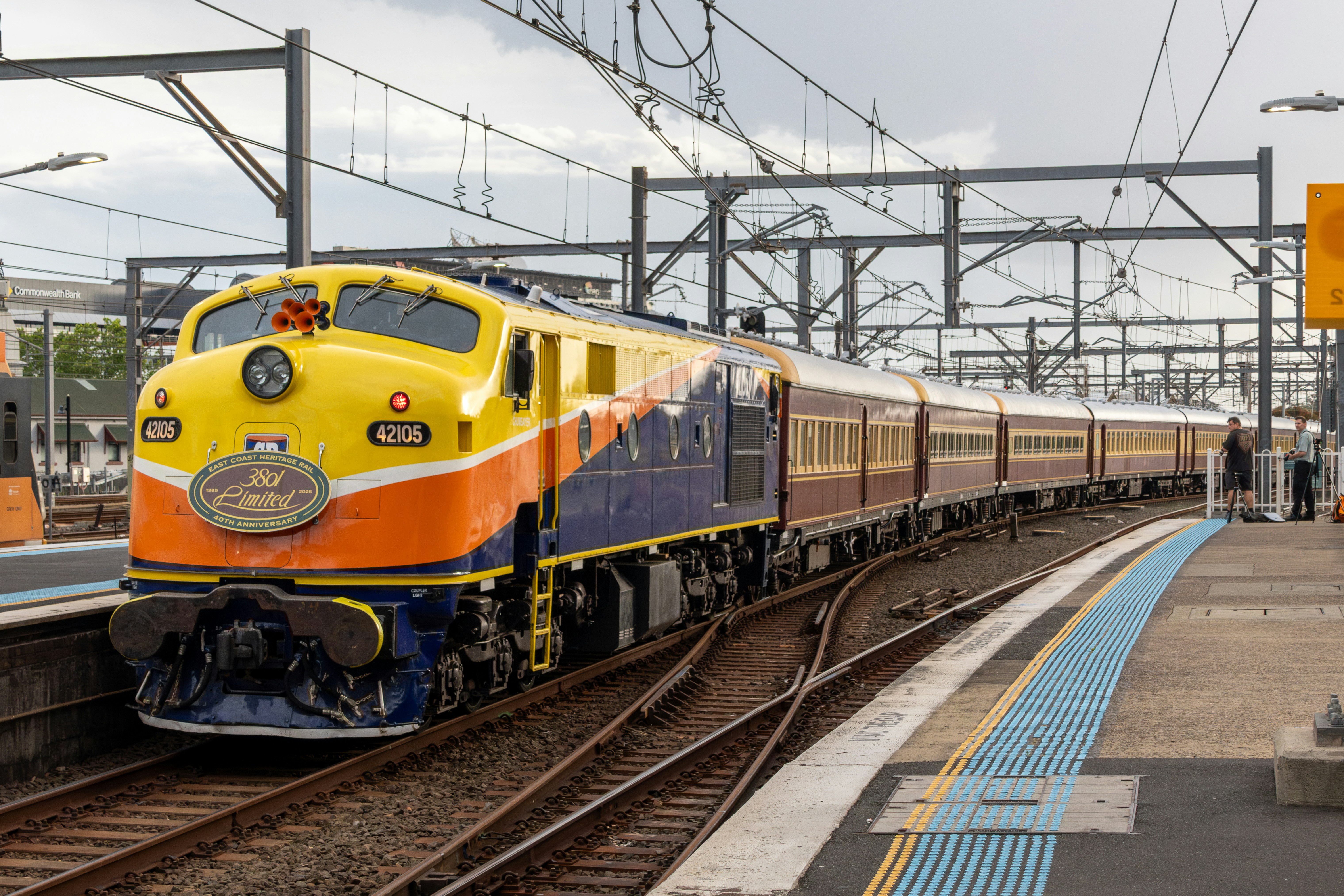 A yellow and blue passenger train at a station platform with overhead wires