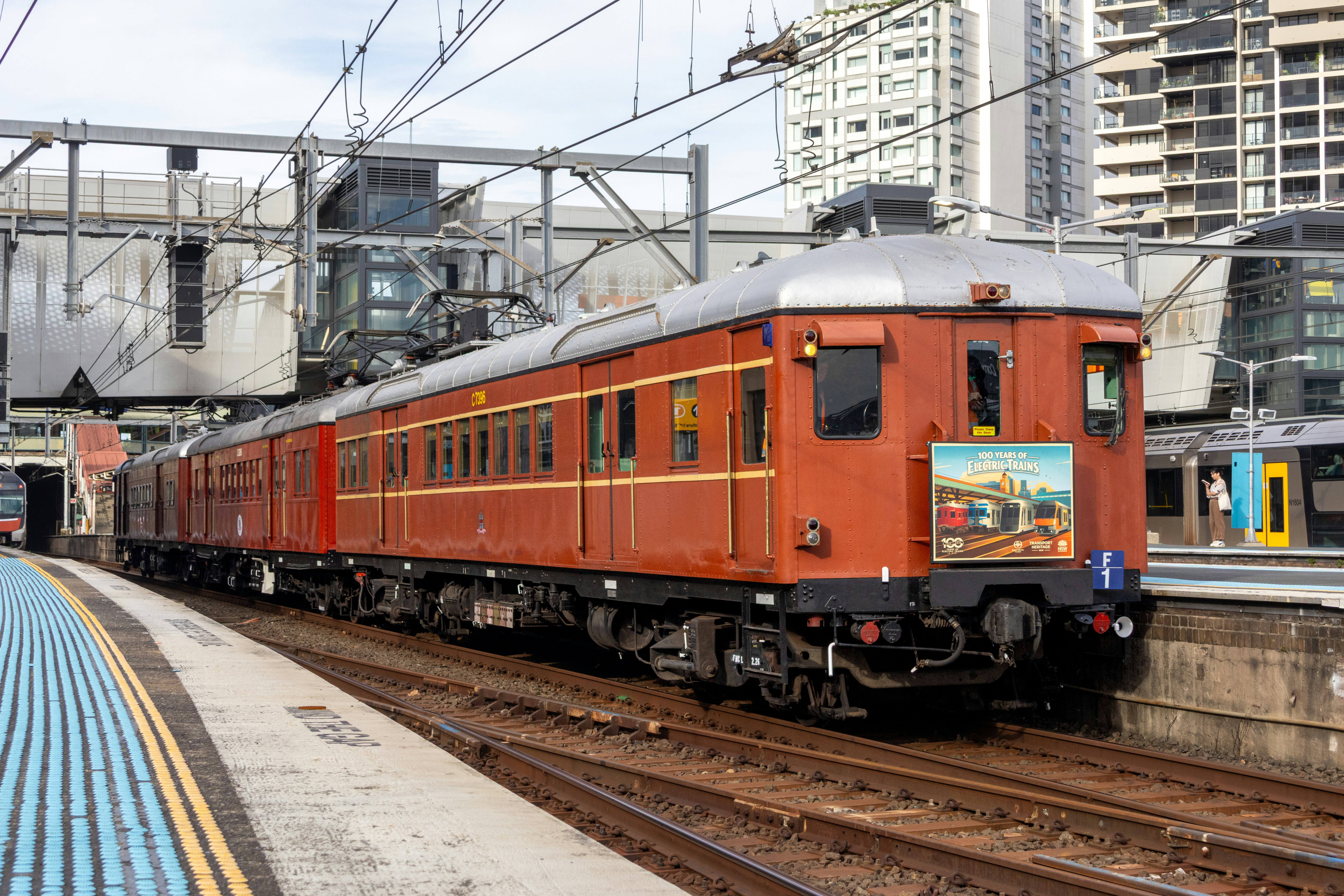 A vintage red electric train at a modern station platform with overhead wires