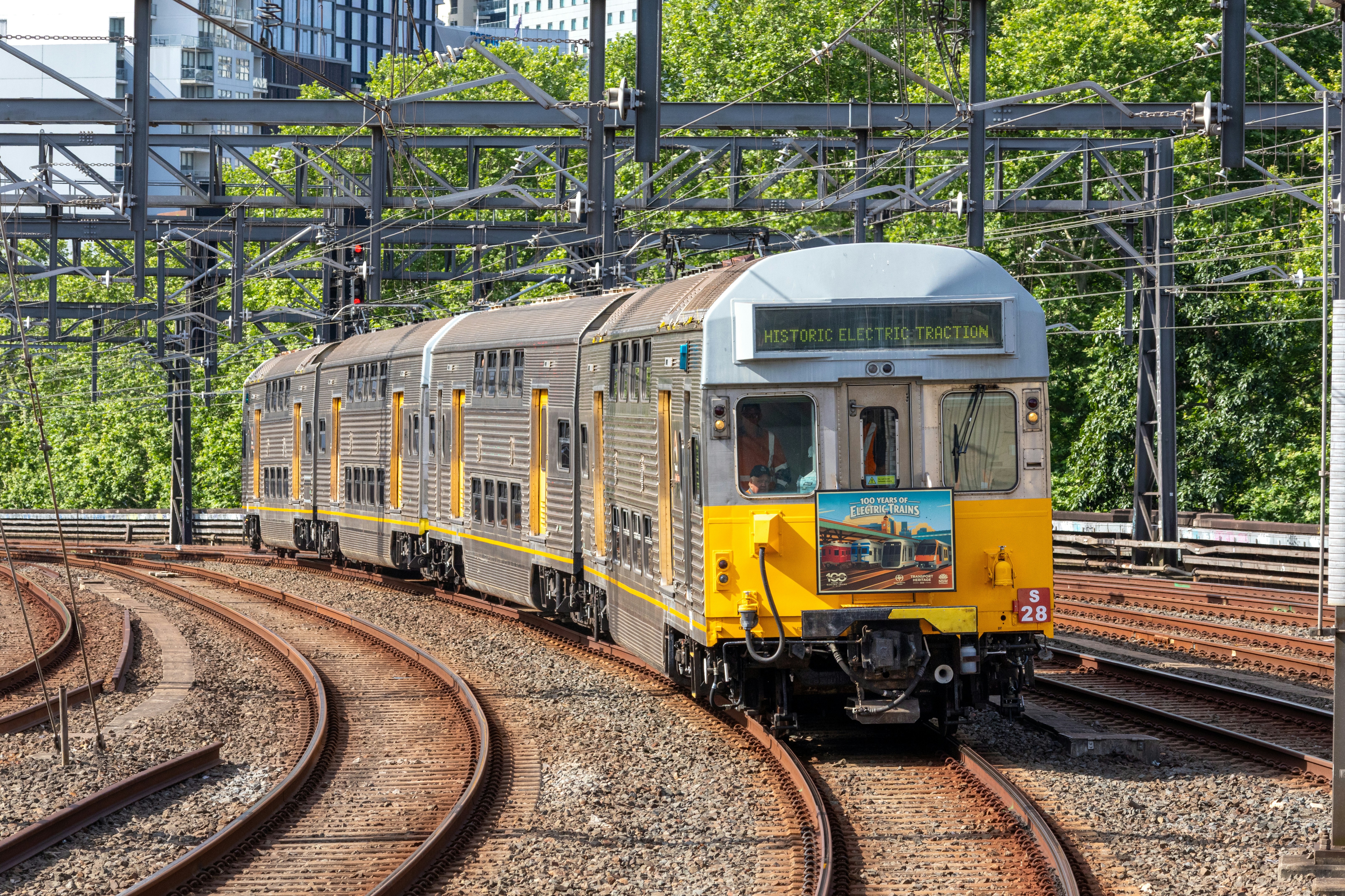 A silver and yellow double-decker train on curved tracks under overhead power lines
