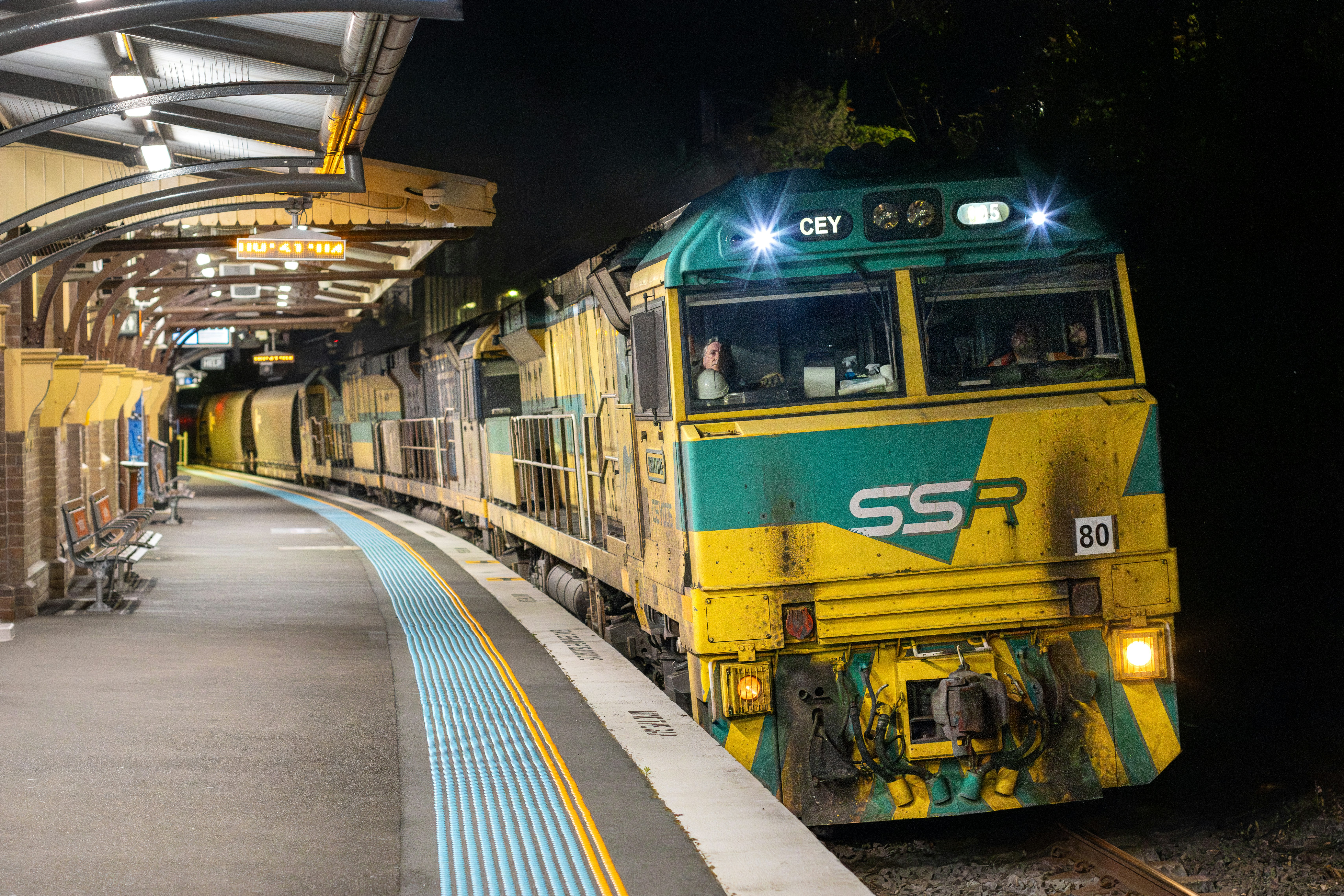 A yellow and green freight train at a railway station platform at night