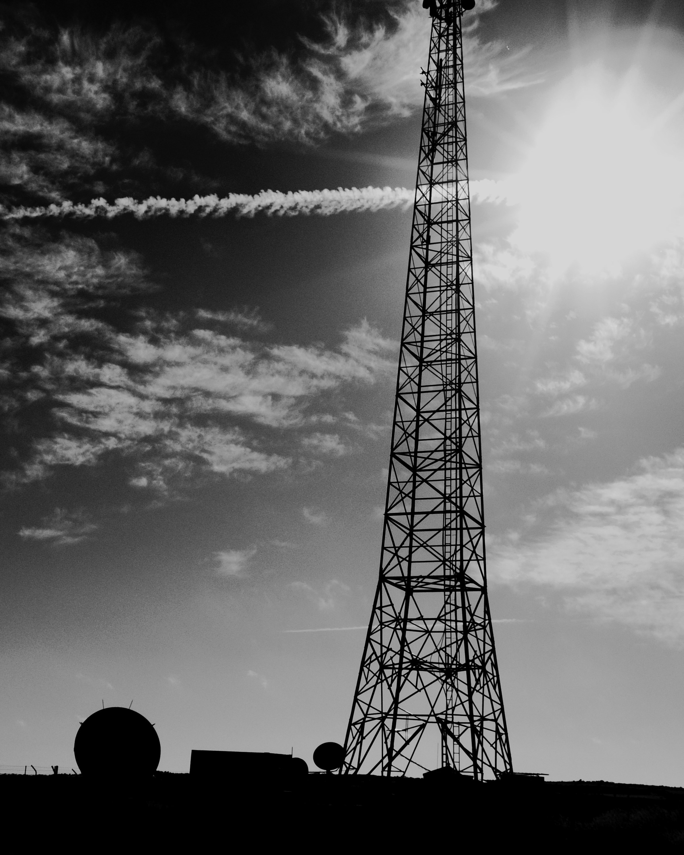A tall lattice communication tower in silhouette against a bright sky with clouds