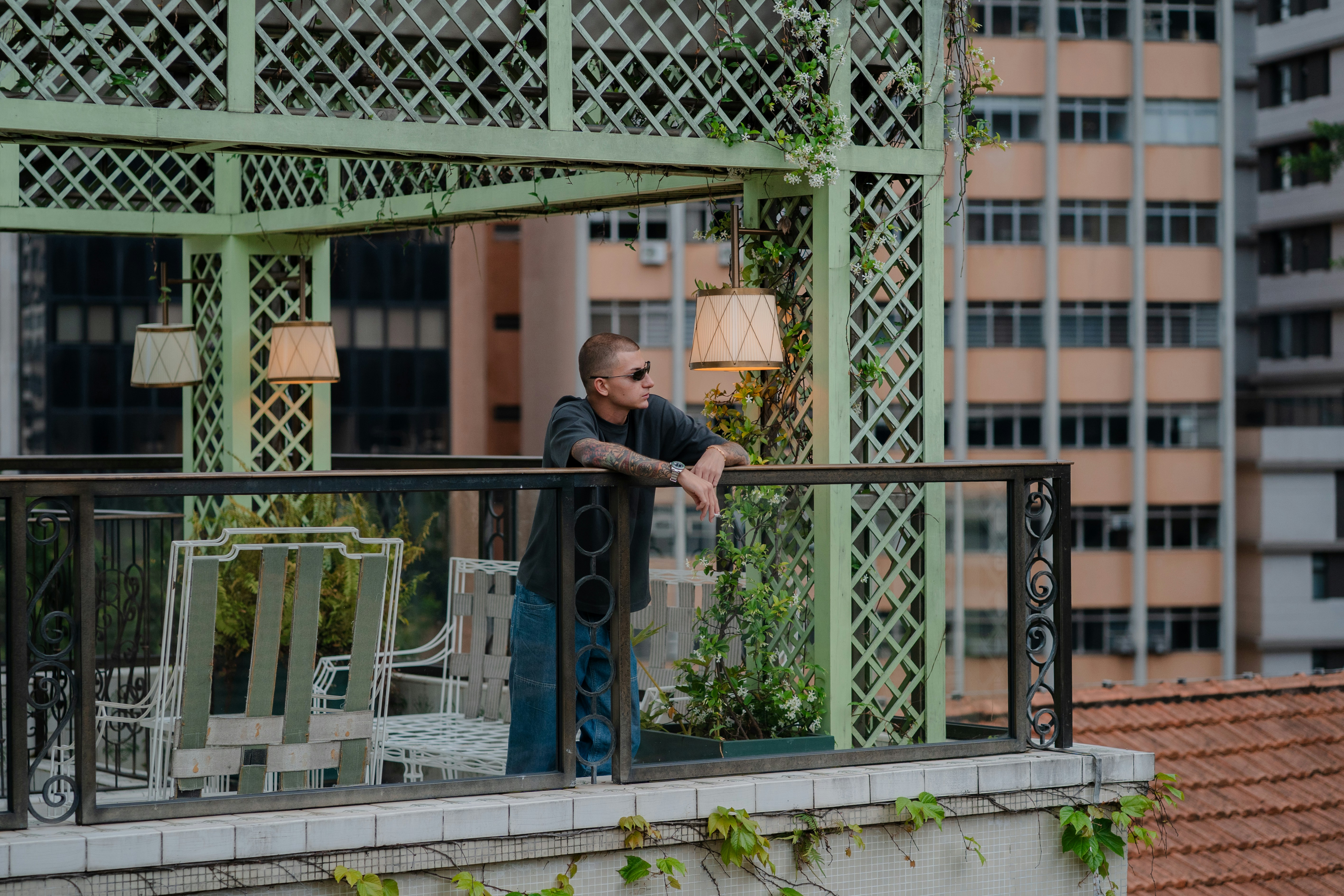 A person with tattoos leaning on a balcony railing in a green trellis gazebo
