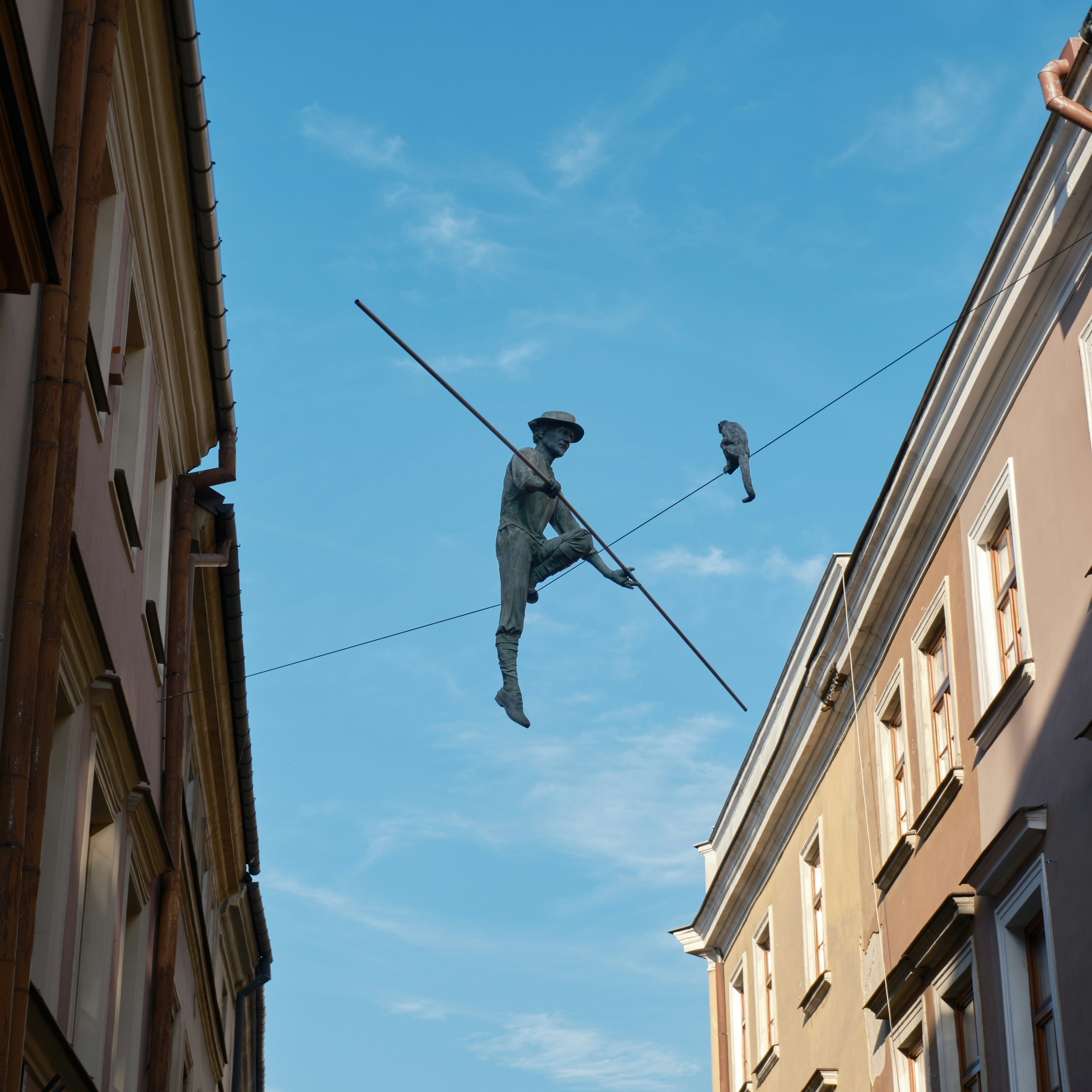 ataxia: A bronze tightrope walker sculpture with a monkey suspended between two city buildings