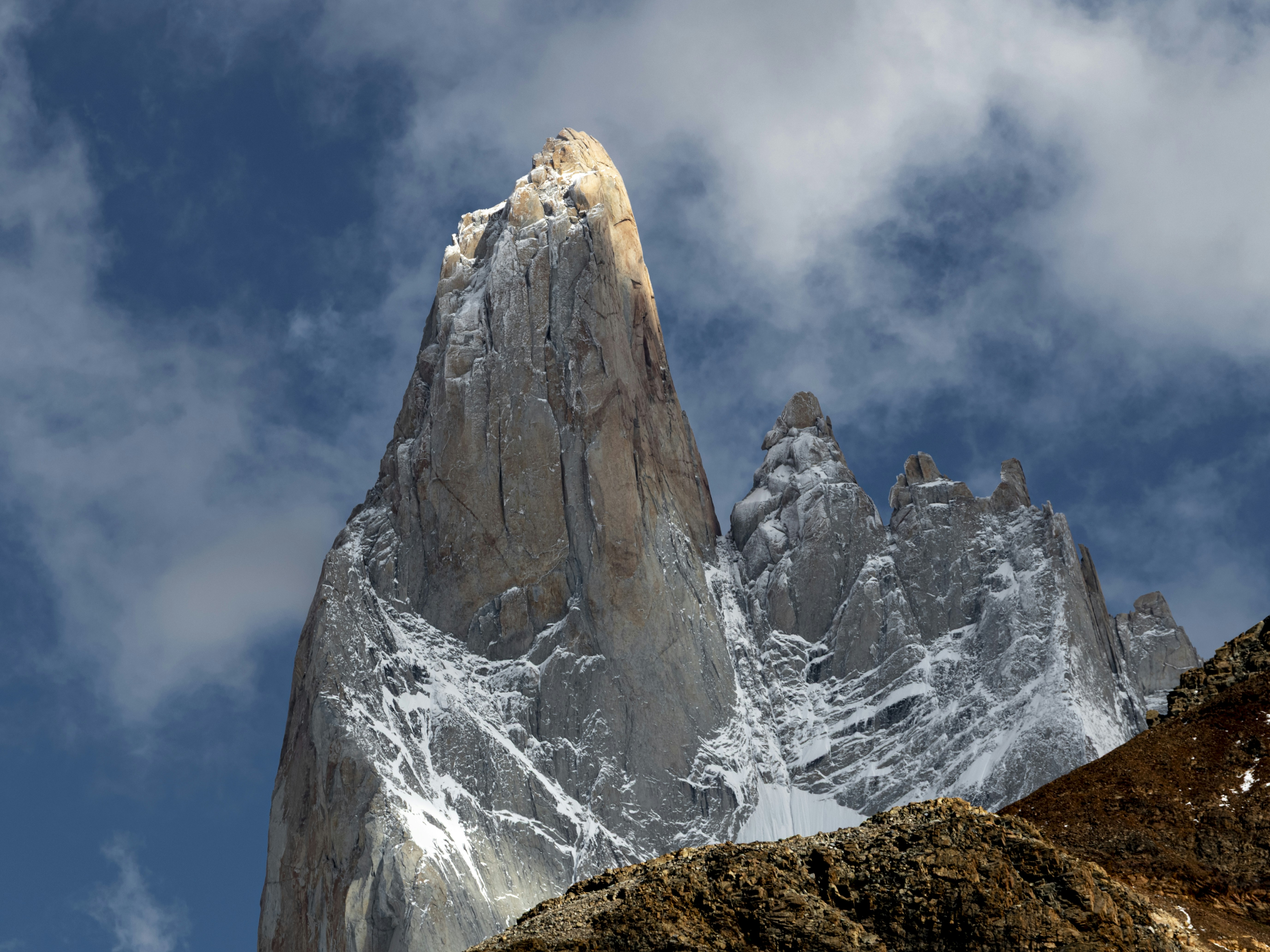 A jagged granite mountain peak with snow patches against a blue cloudy sky