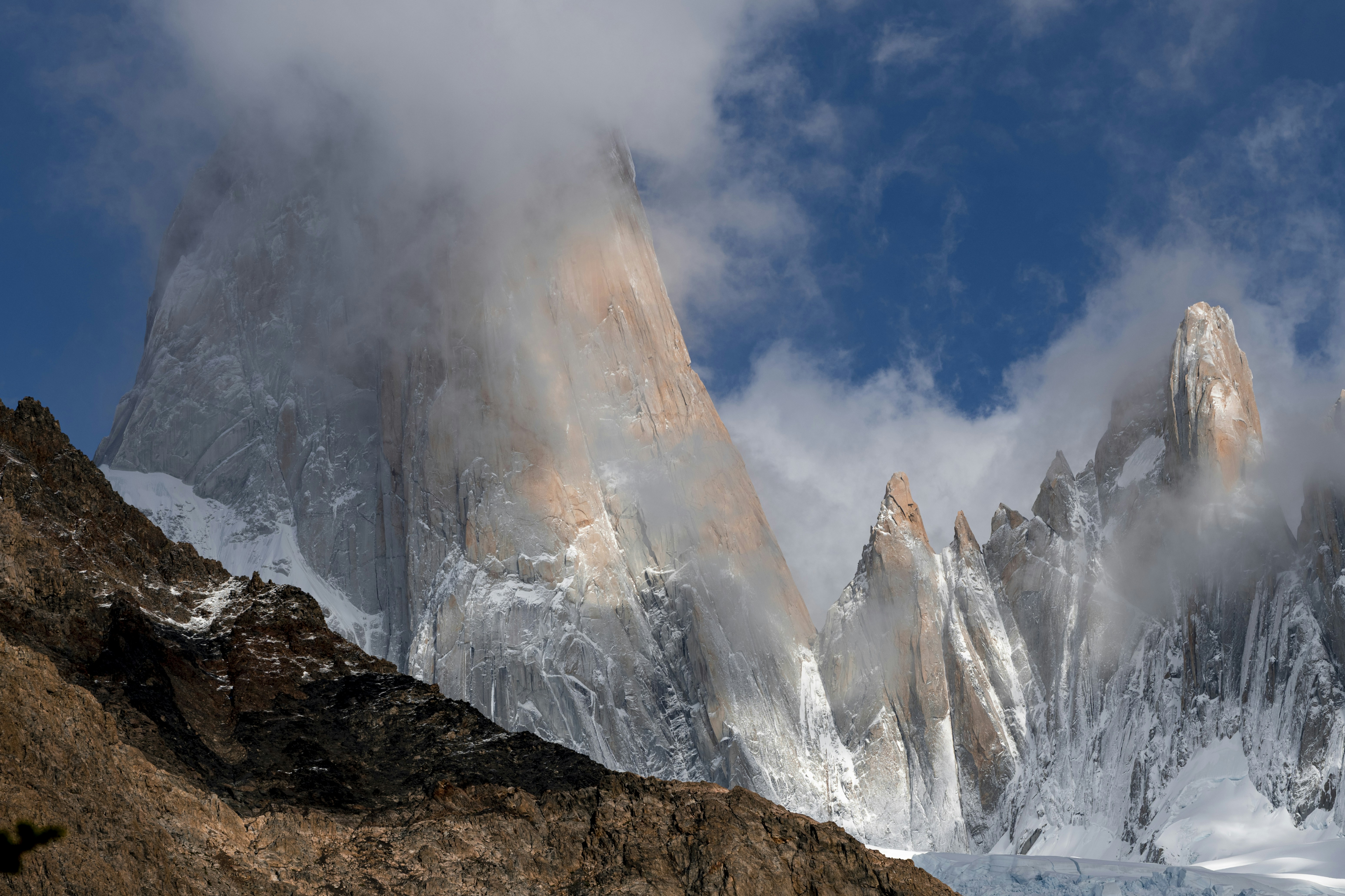 Jagged snow-covered mountain peaks partially obscured by clouds under a deep blue sky