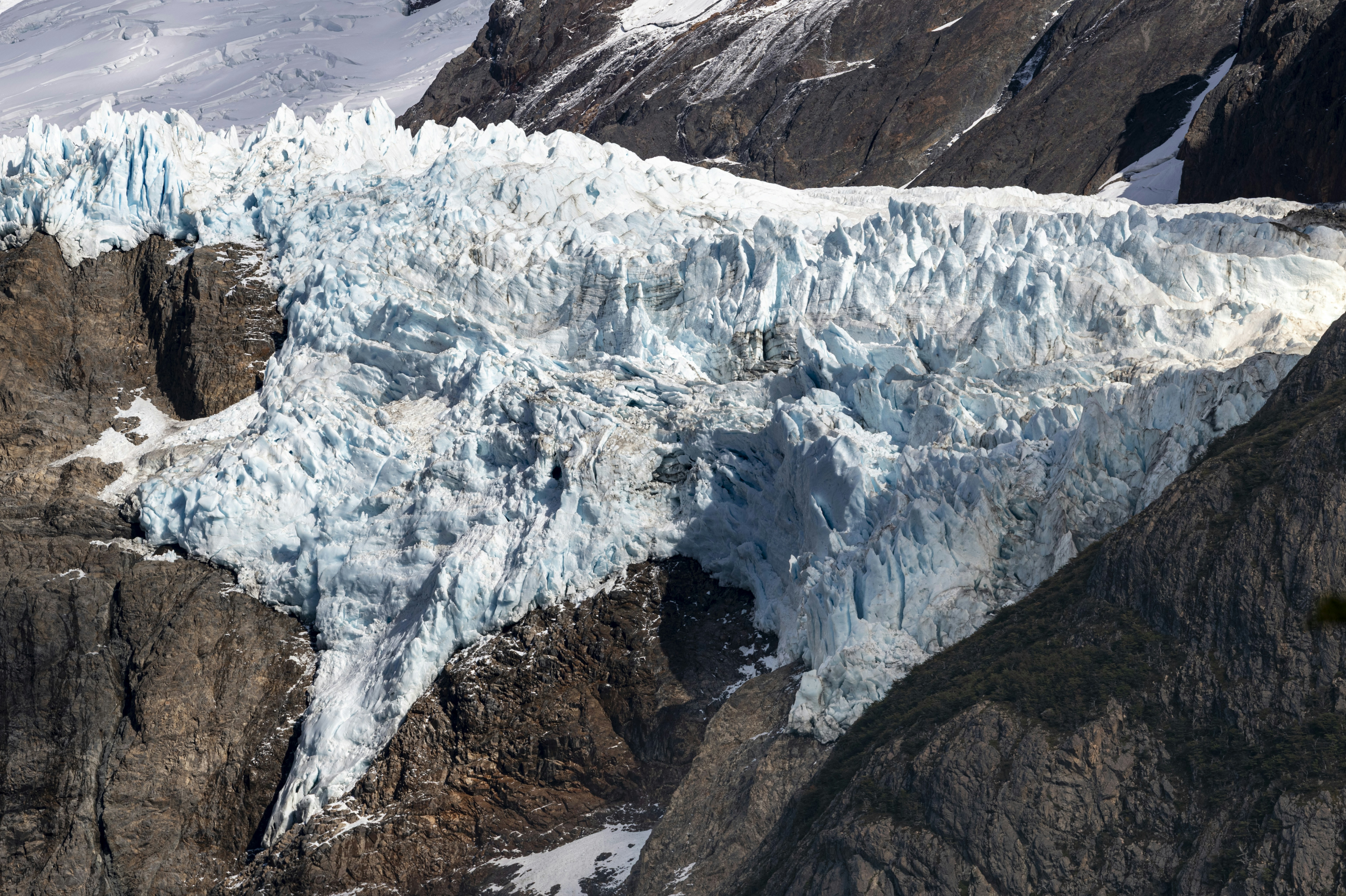 A jagged blue glacier flowing down between steep dark rocky mountain slopes