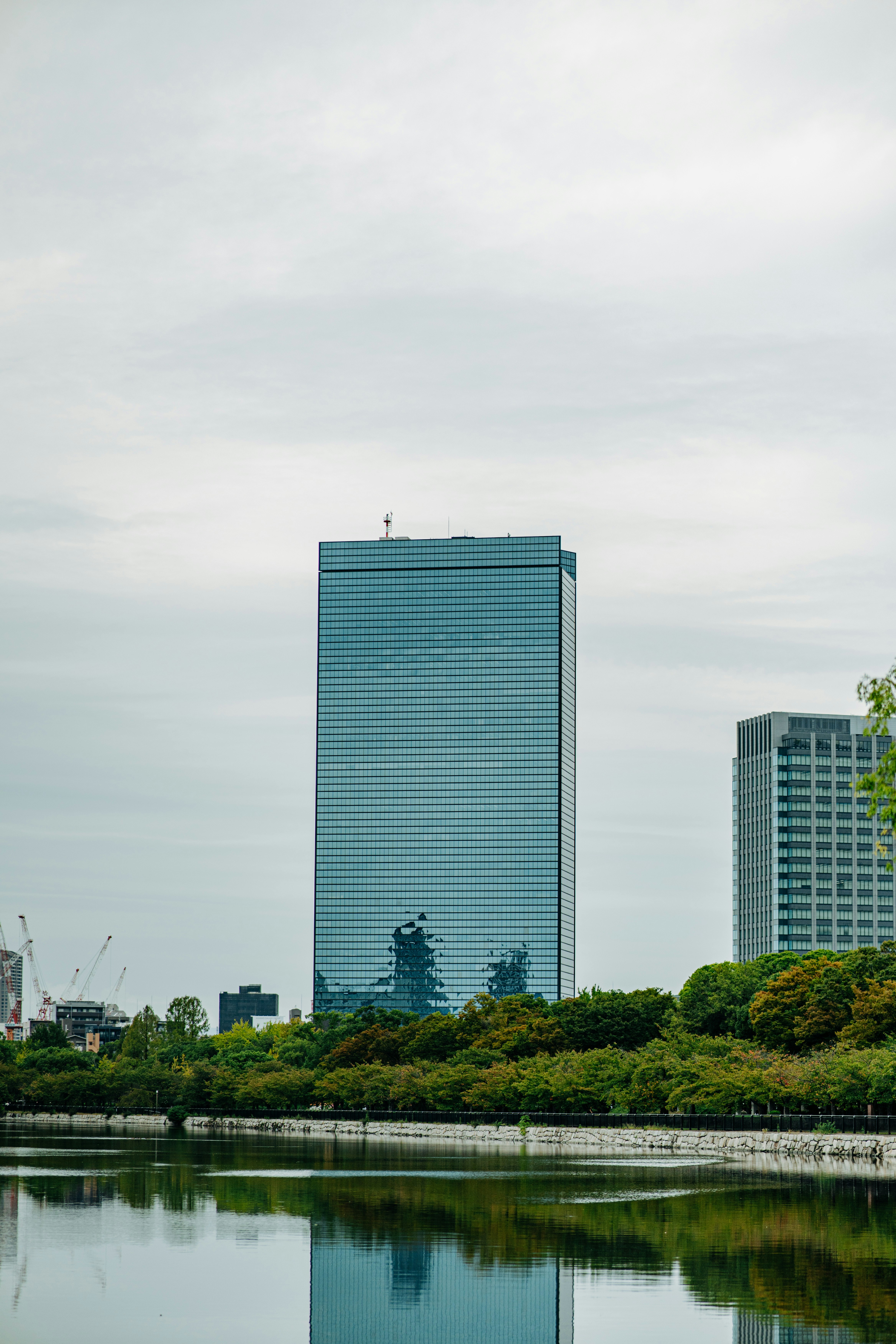 A tall glass skyscraper reflecting the sky above a calm lake and trees