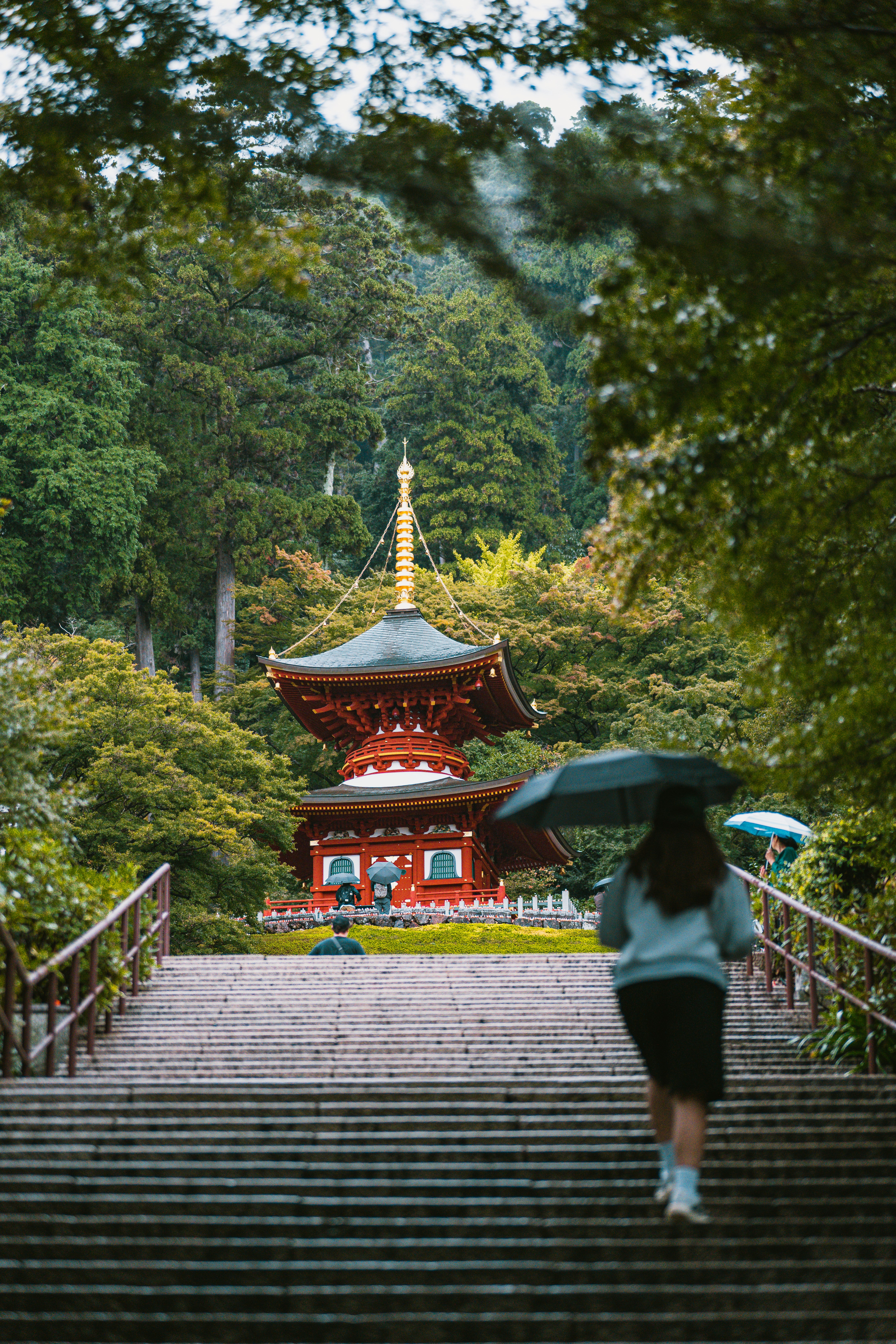 A person with an umbrella walking up stone stairs toward a red pagoda