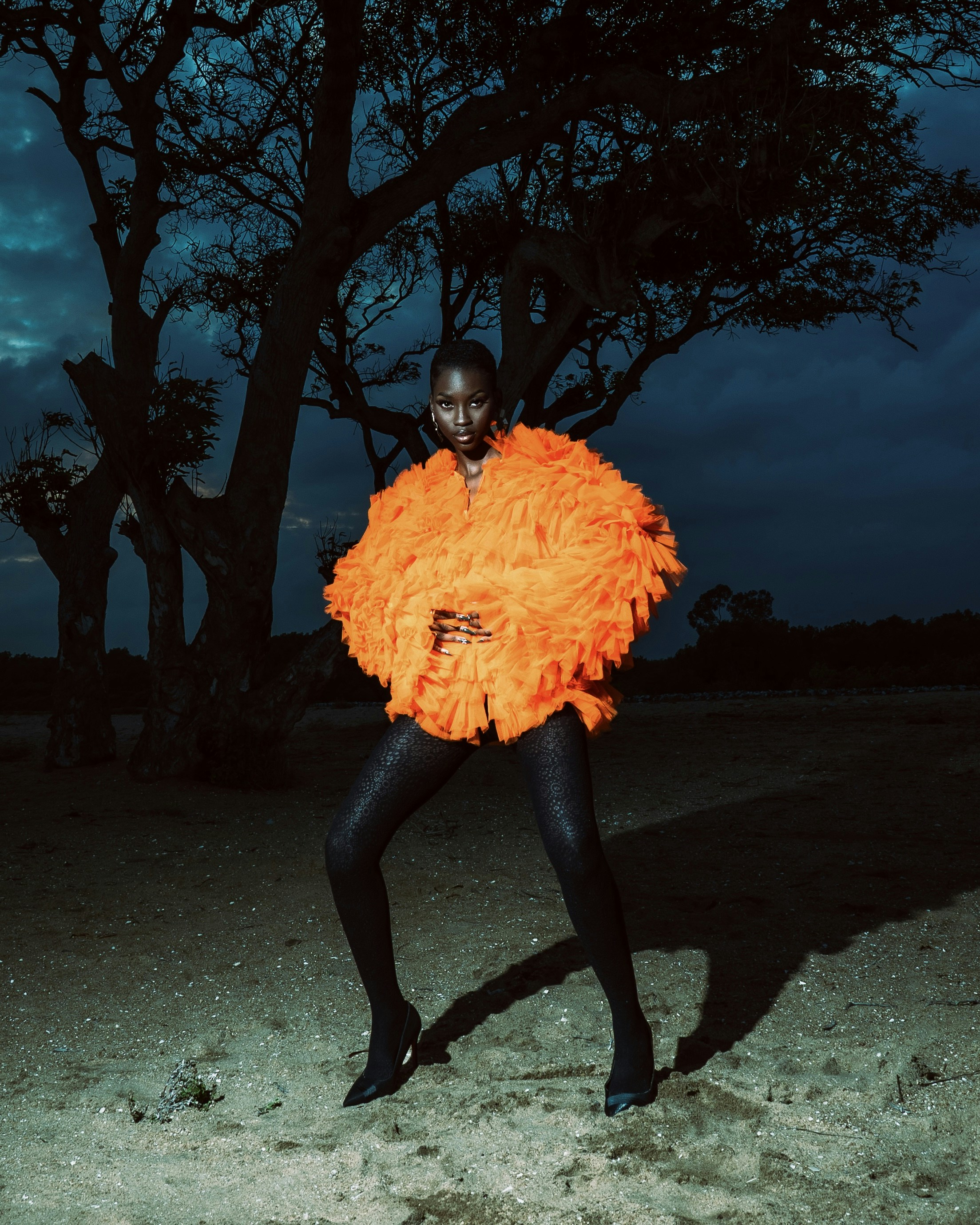 A person in a bright orange ruffled jacket and black leggings standing before a dark tree at night