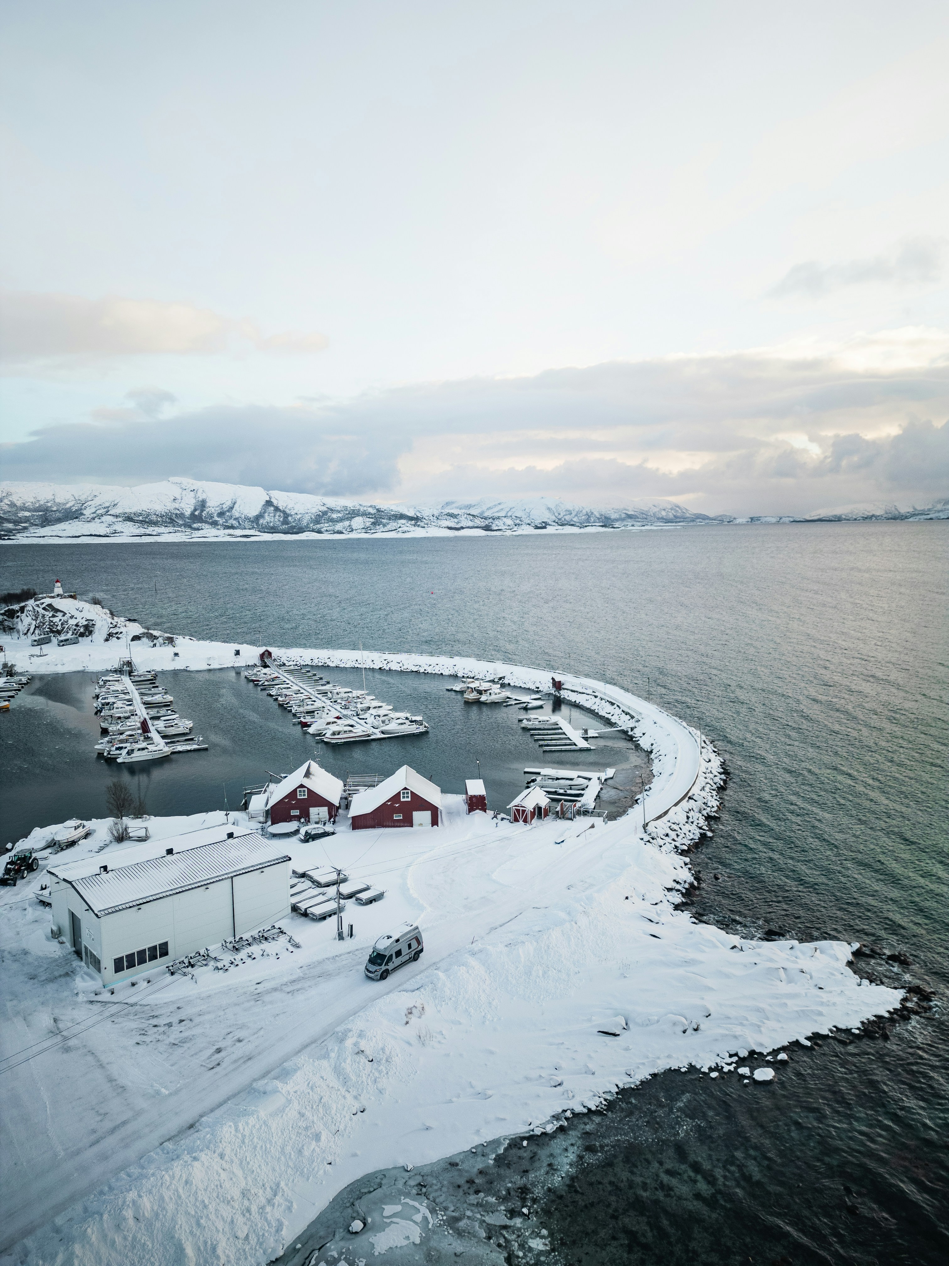 A camper van on a snowy road beside a marina with red buildings