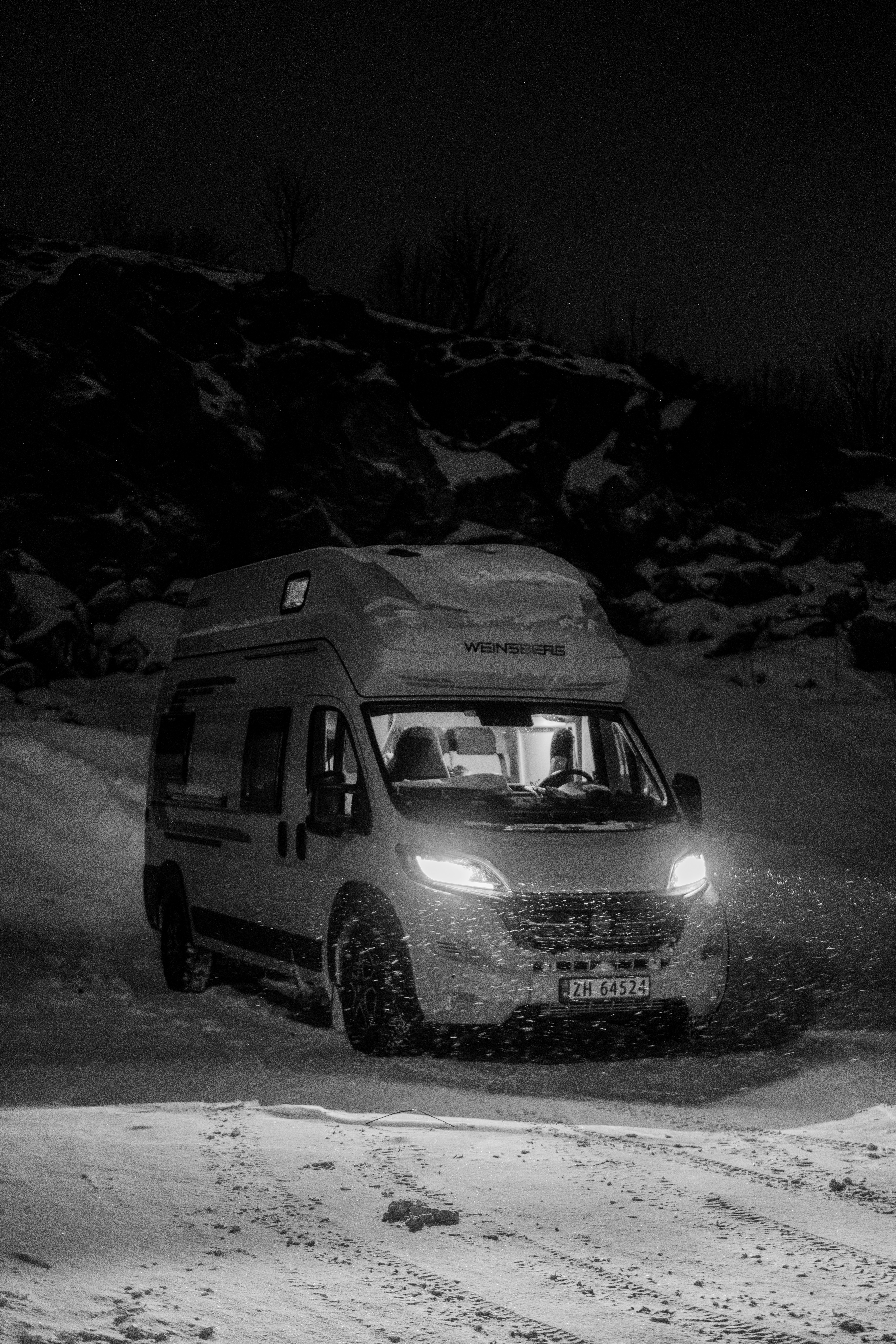 A camper van with illuminated headlights parked on a snowy slope at night