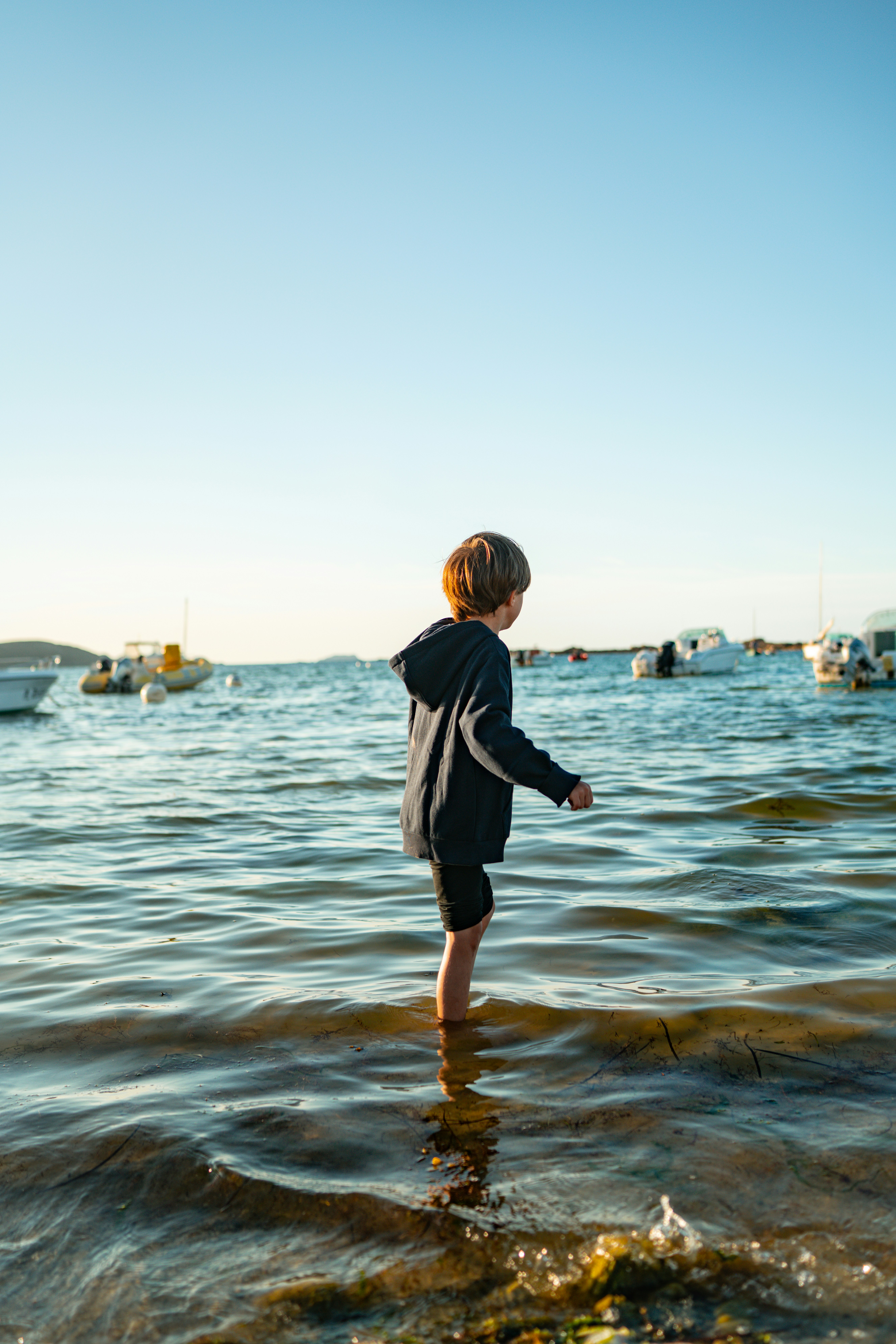 A child in a dark hoodie wading in shallow water near moored boats - 相关推荐
