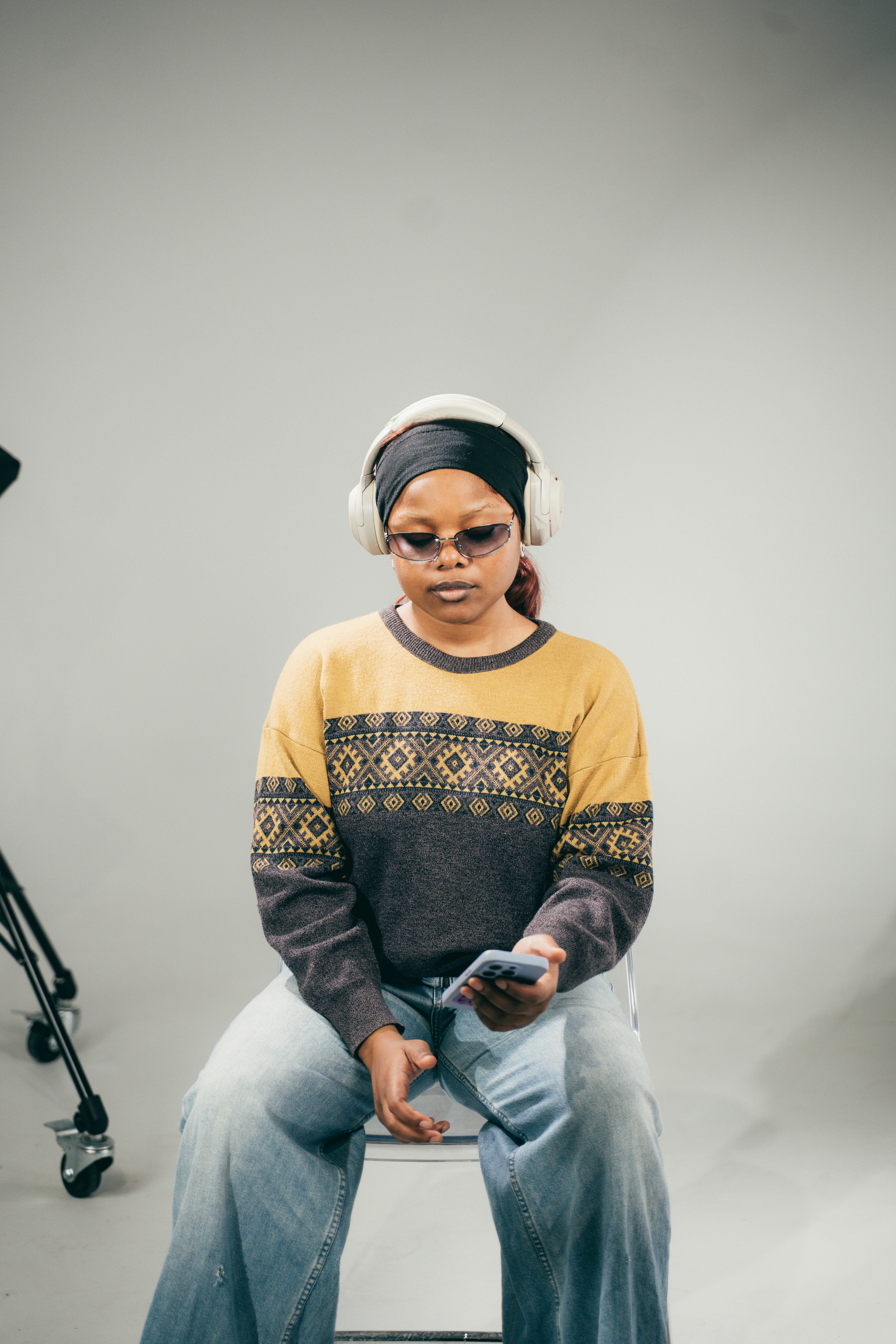 A person wearing headphones and sunglasses looking at a smartphone while sitting on a chair