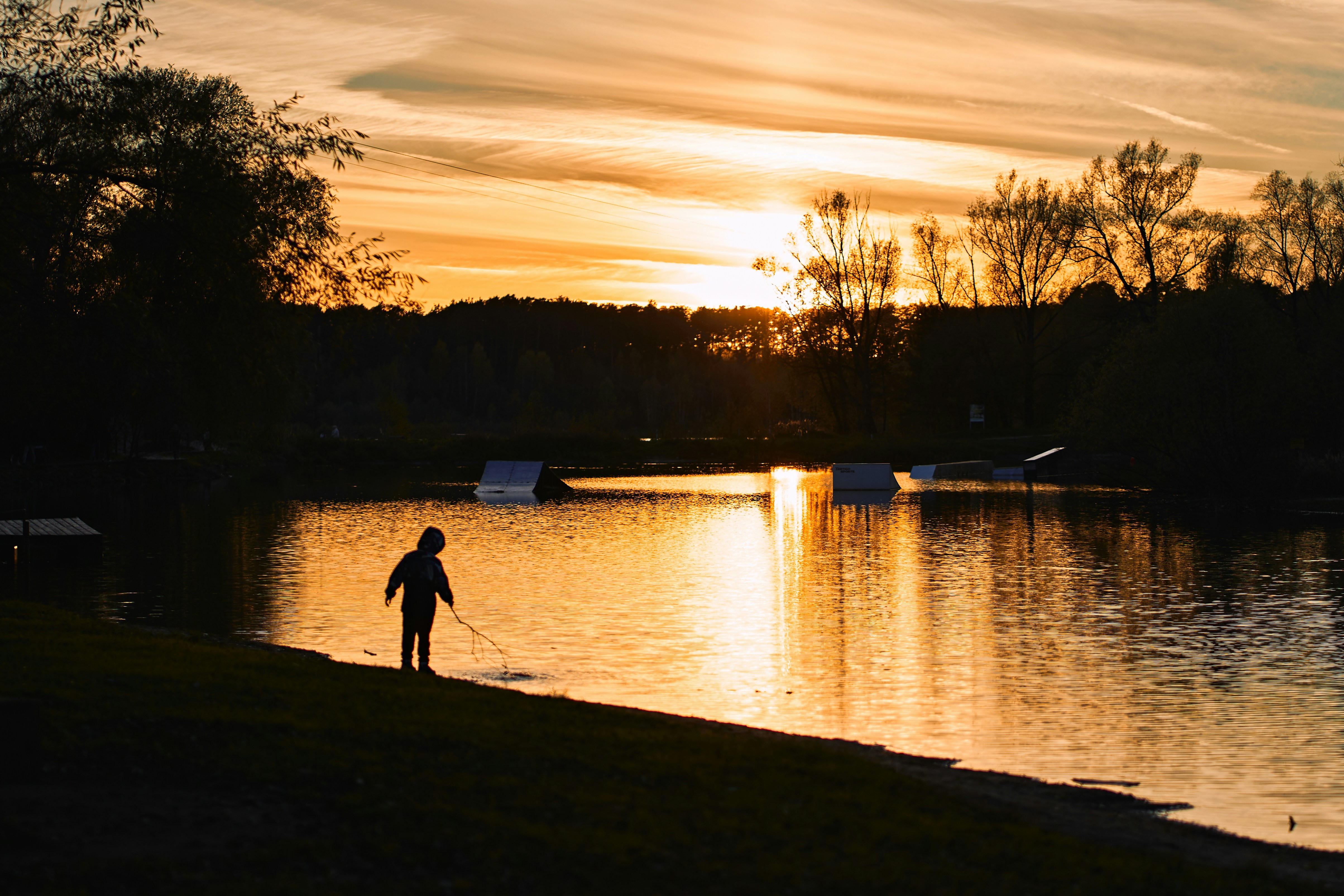 A child in silhouette holding a stick by a lake during golden hour
