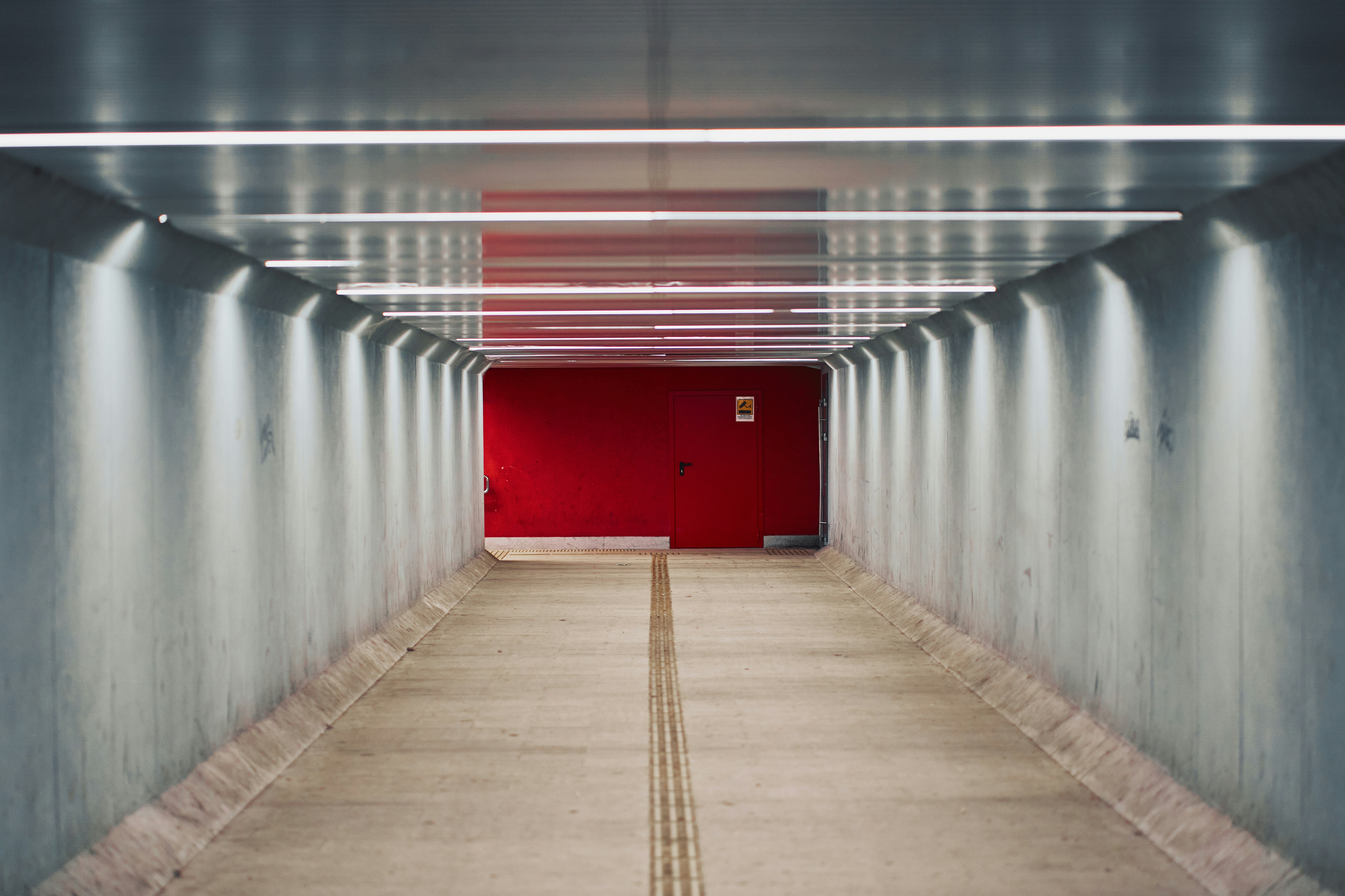 A concrete pedestrian tunnel with bright overhead lights leading to a vibrant red door