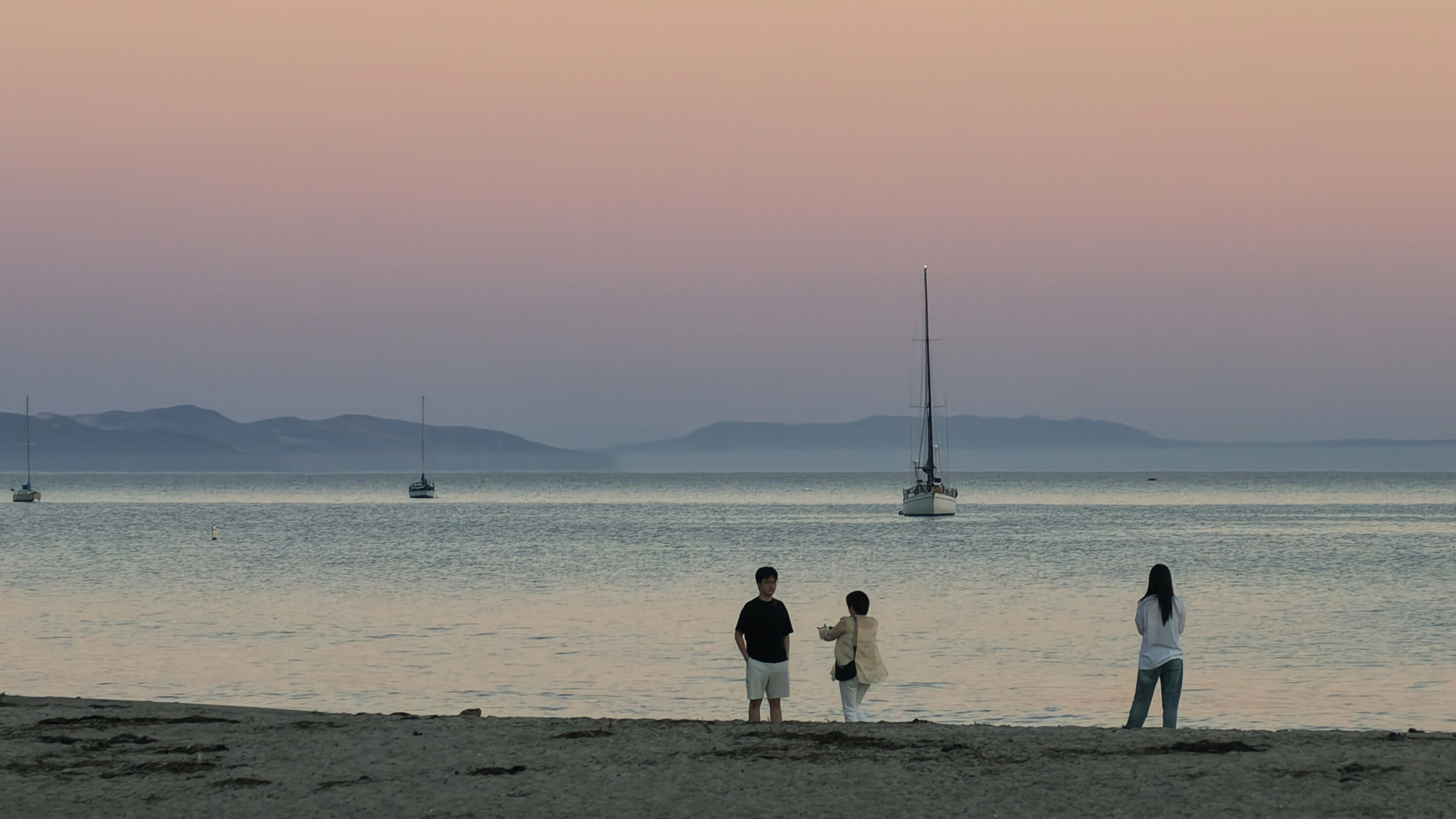 Tres personas en una playa de arena bajo el sol bajo con veleros en el agua