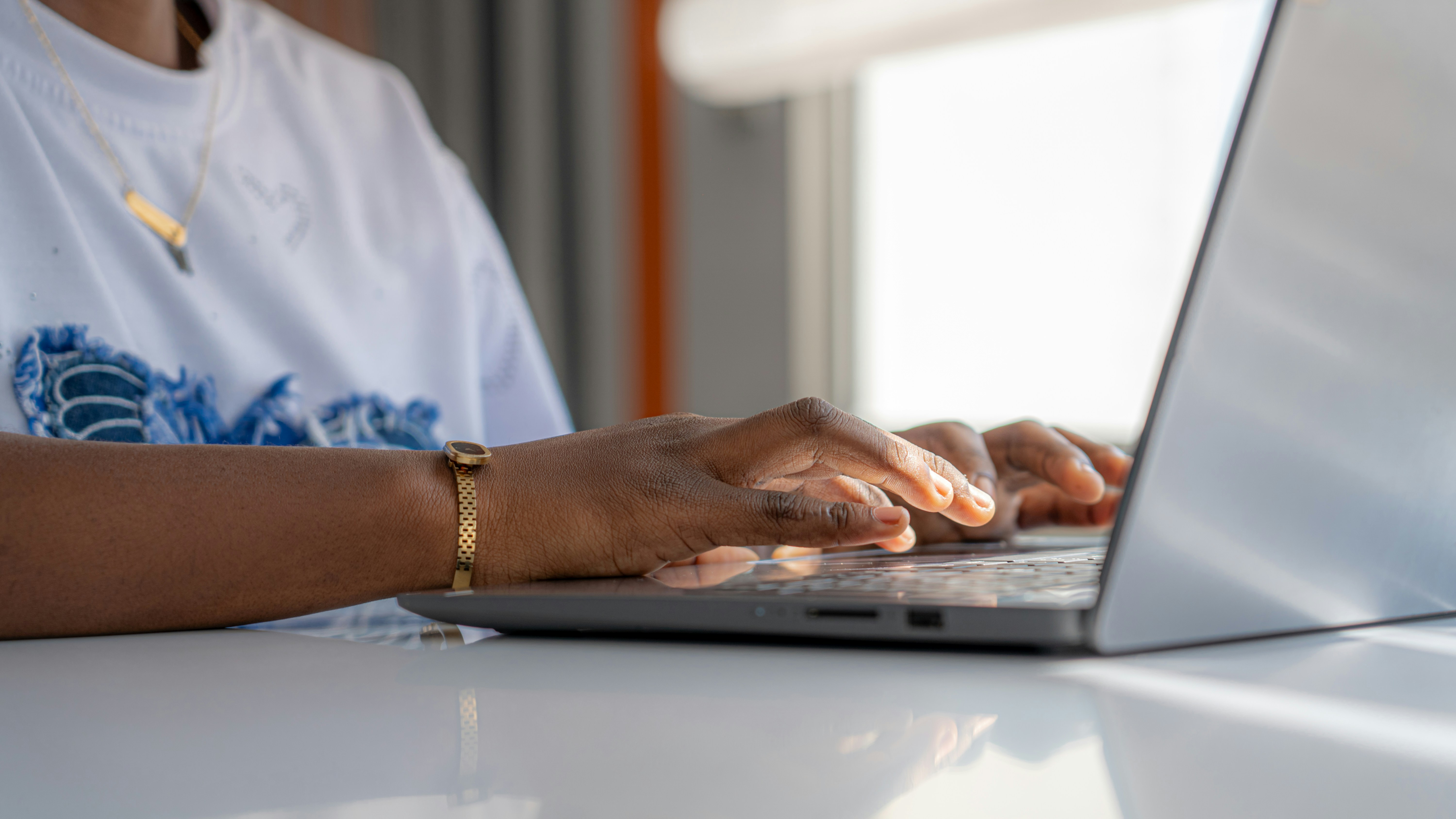 A person typing on a laptop at a white desk in a sunlit room