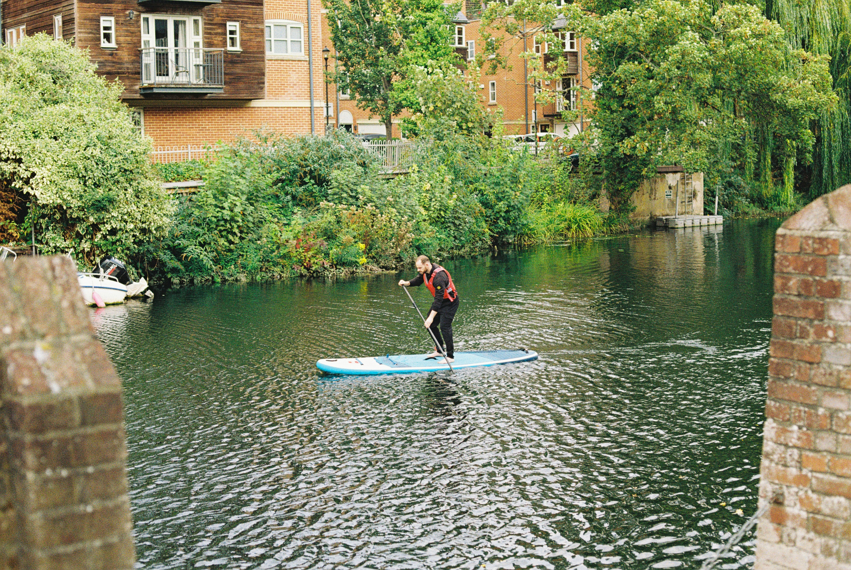 A man in a life vest paddleboarding on a calm river near brick buildings
