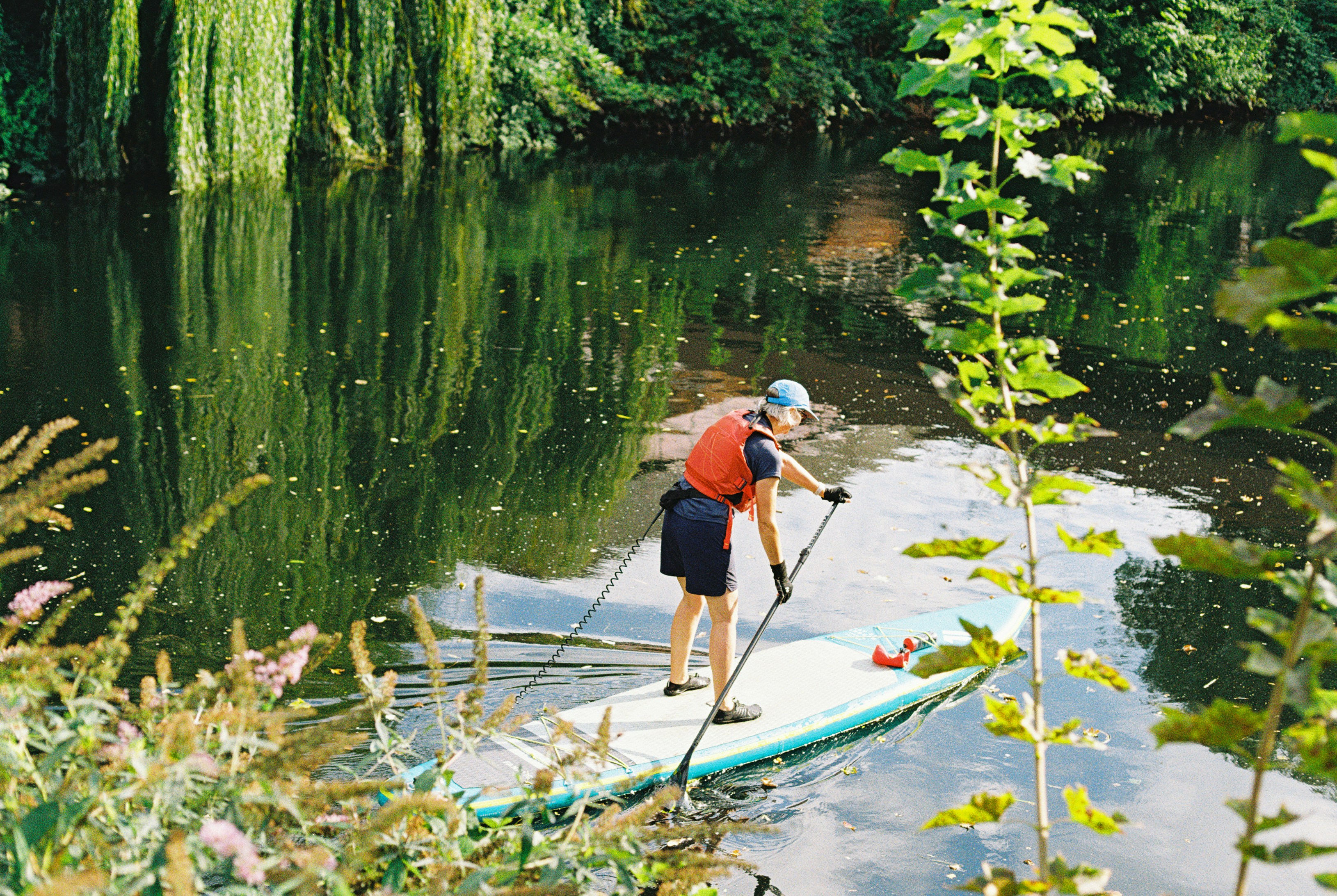 A person in a life vest paddleboarding on a calm river lined with willow trees