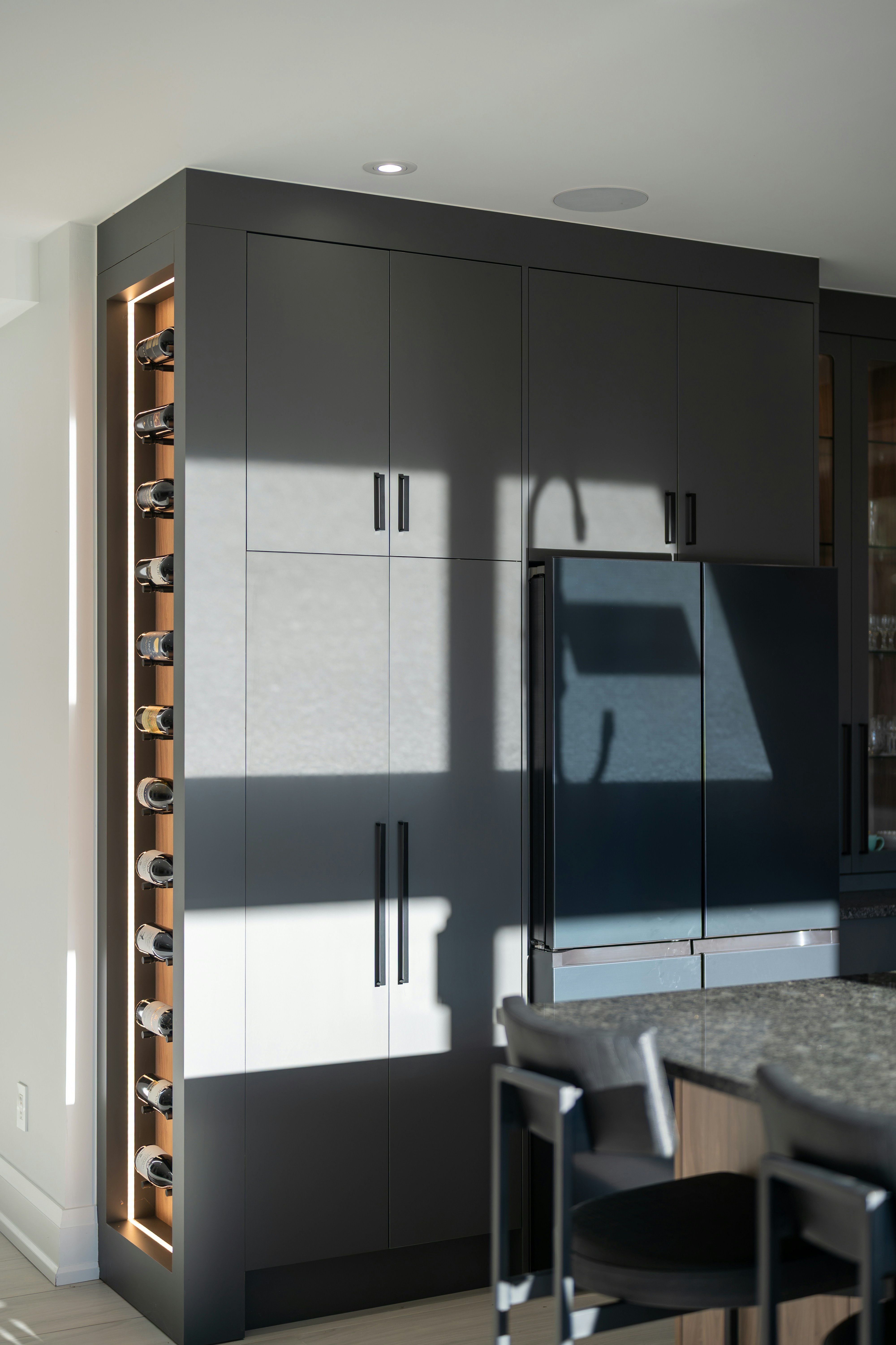 A modern kitchen with dark grey cabinets, a built-in wine rack, and a refrigerator in bright sunlight