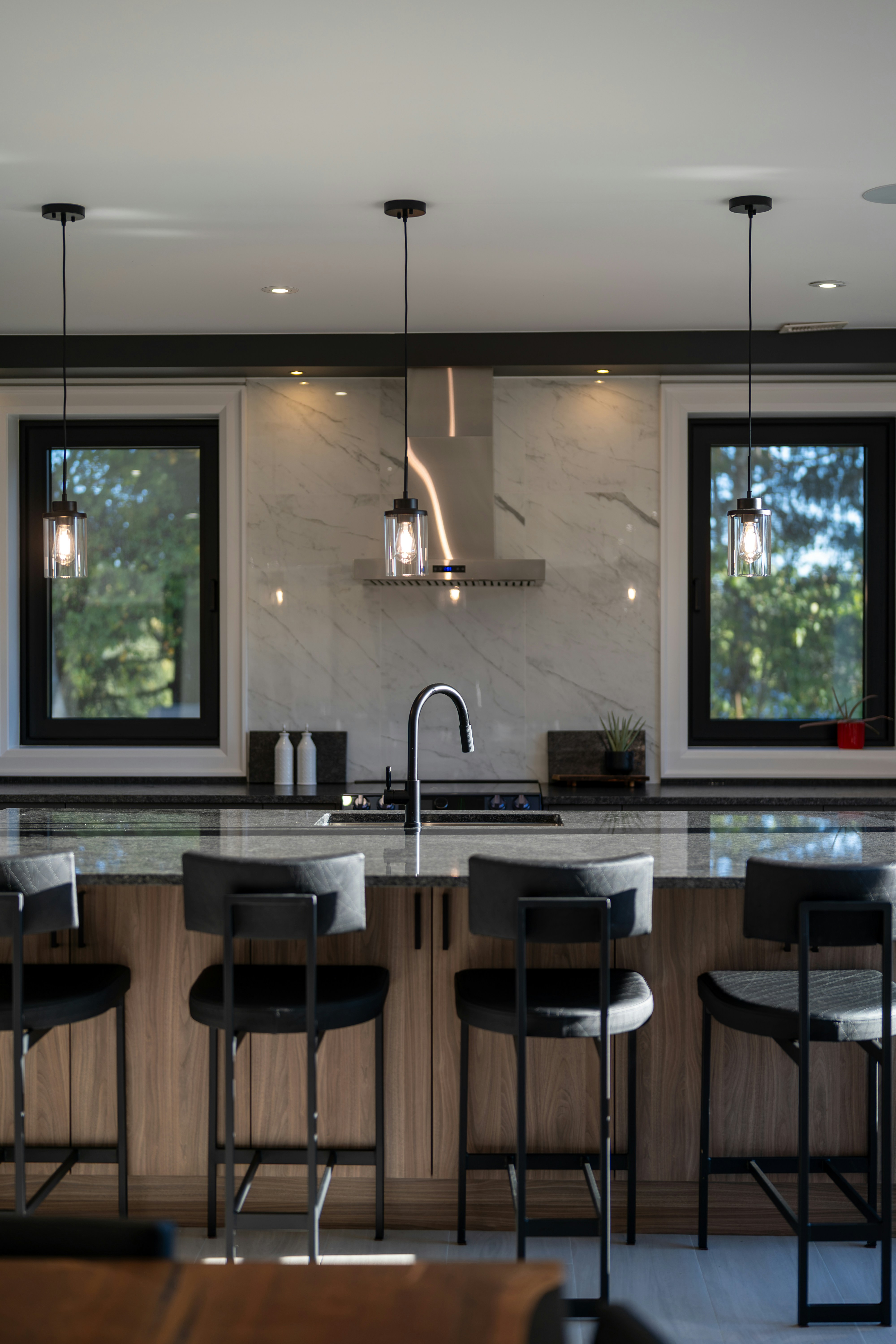 A kitchen island with four black stools in front of a marble backsplash and windows