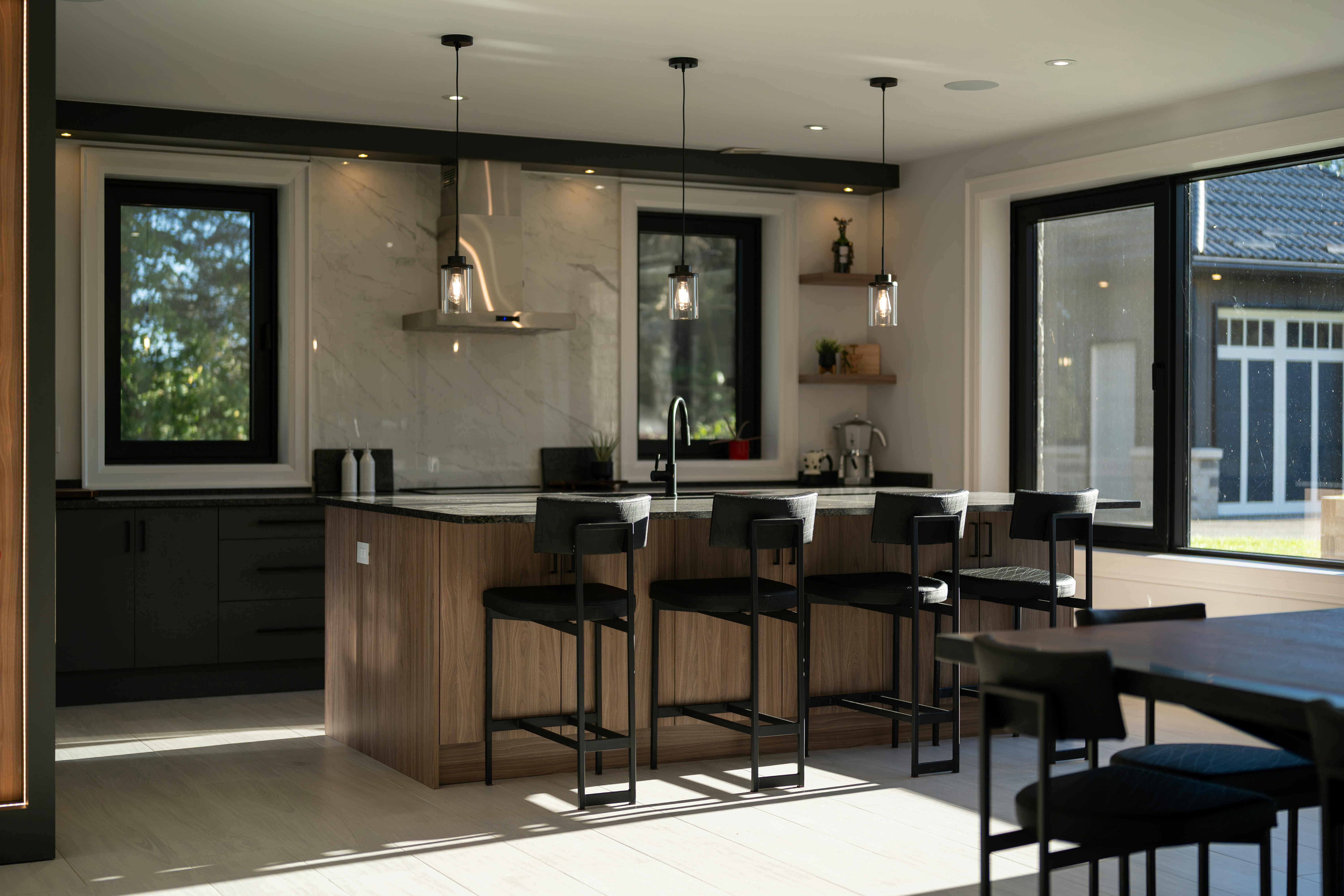 A modern kitchen with a wooden island, black bar stools, and marble backsplash in bright sunlight