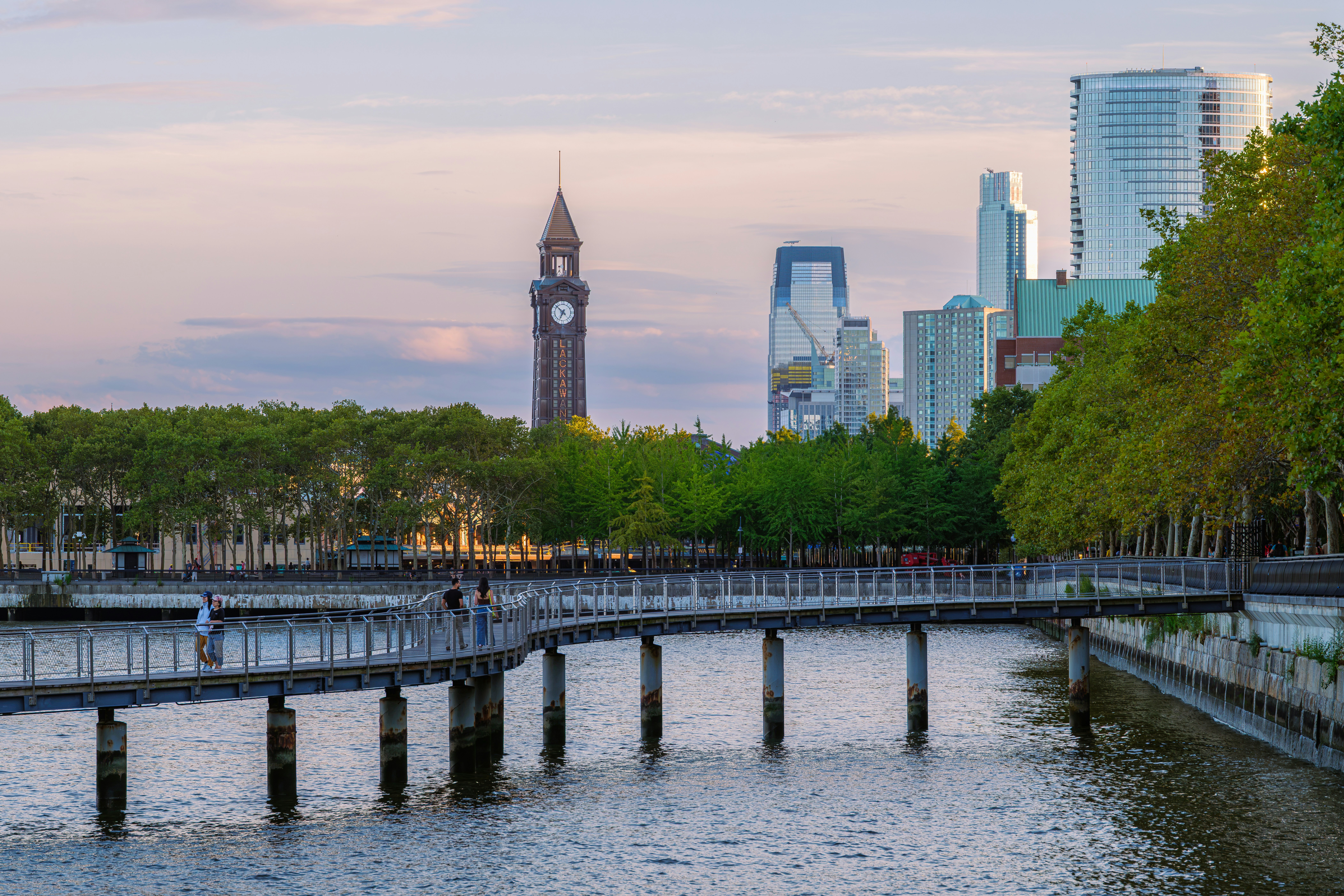 Curving waterfront walkway leads toward the Lackawanna clock tower and modern Hoboken skyline in soft evening light, New