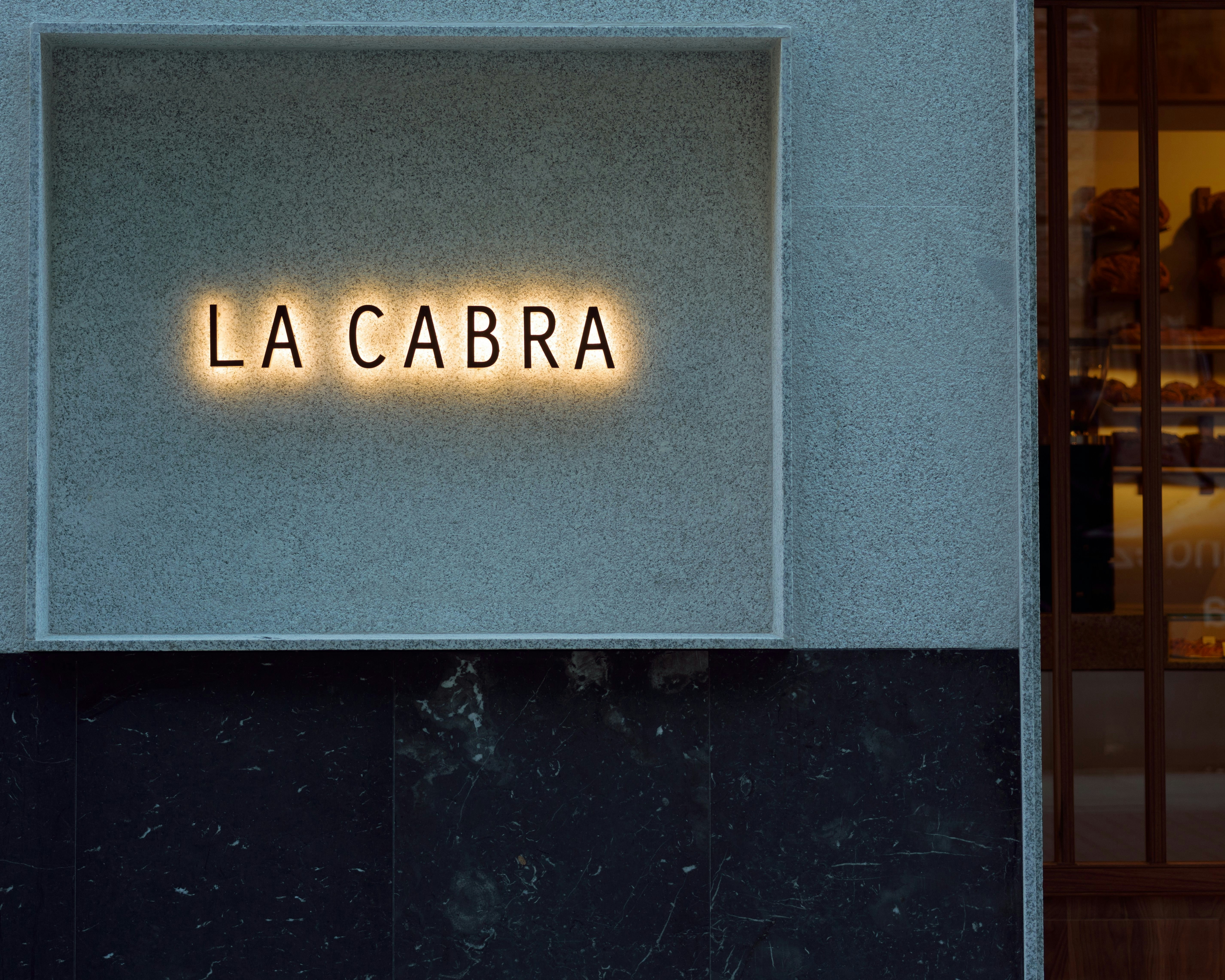 A glowing sign reading LA CABRA on a textured grey wall above dark marble panels