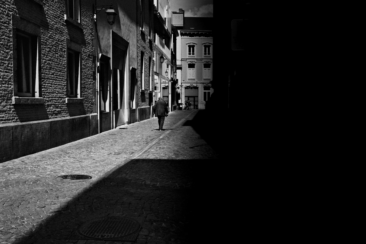 A person walking with a cane down a narrow cobblestone alley in high contrast light