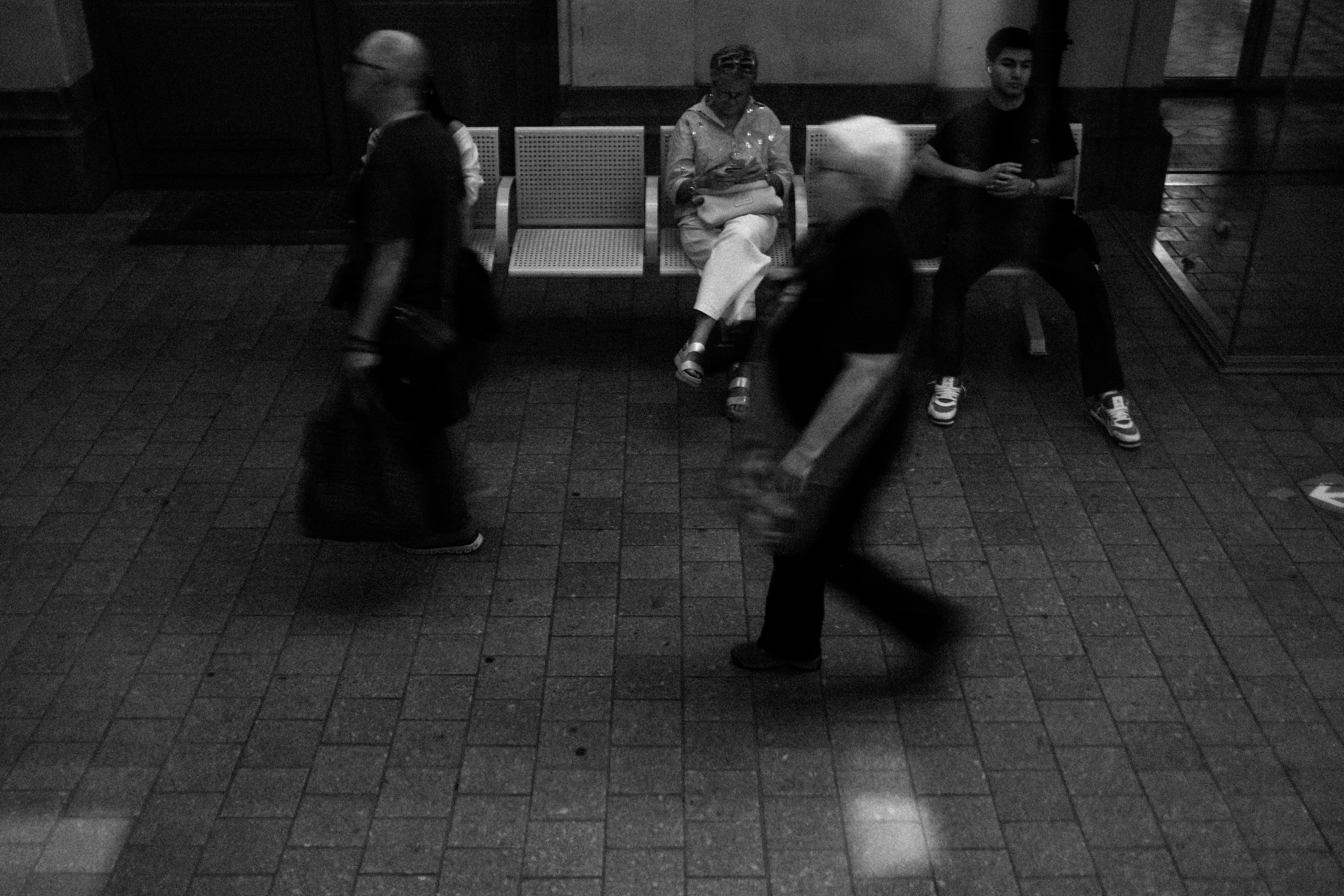 People walking past others seated on metal chairs in a black and white station