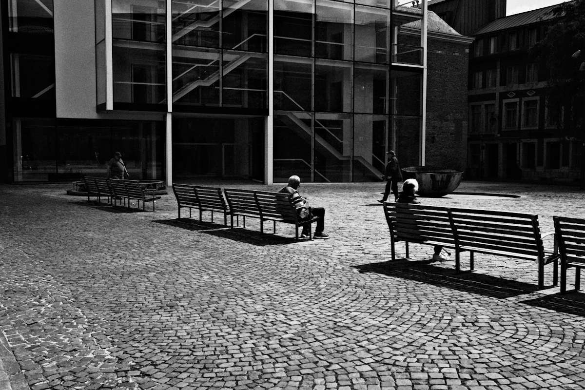 People sitting on wooden benches in a cobblestone square before a modern glass building