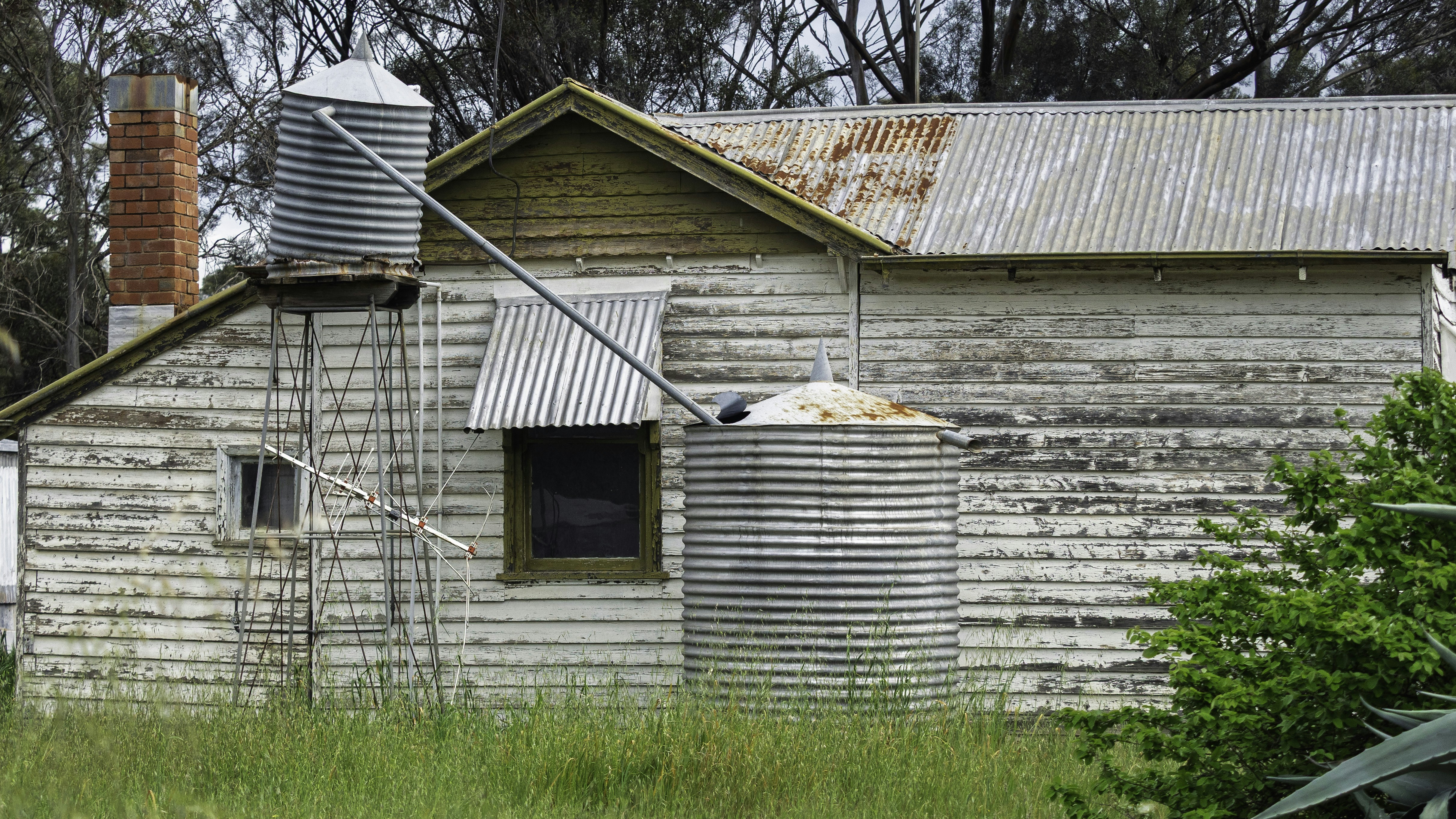 A weathered white wooden cottage with corrugated metal water tanks and a brick chimney