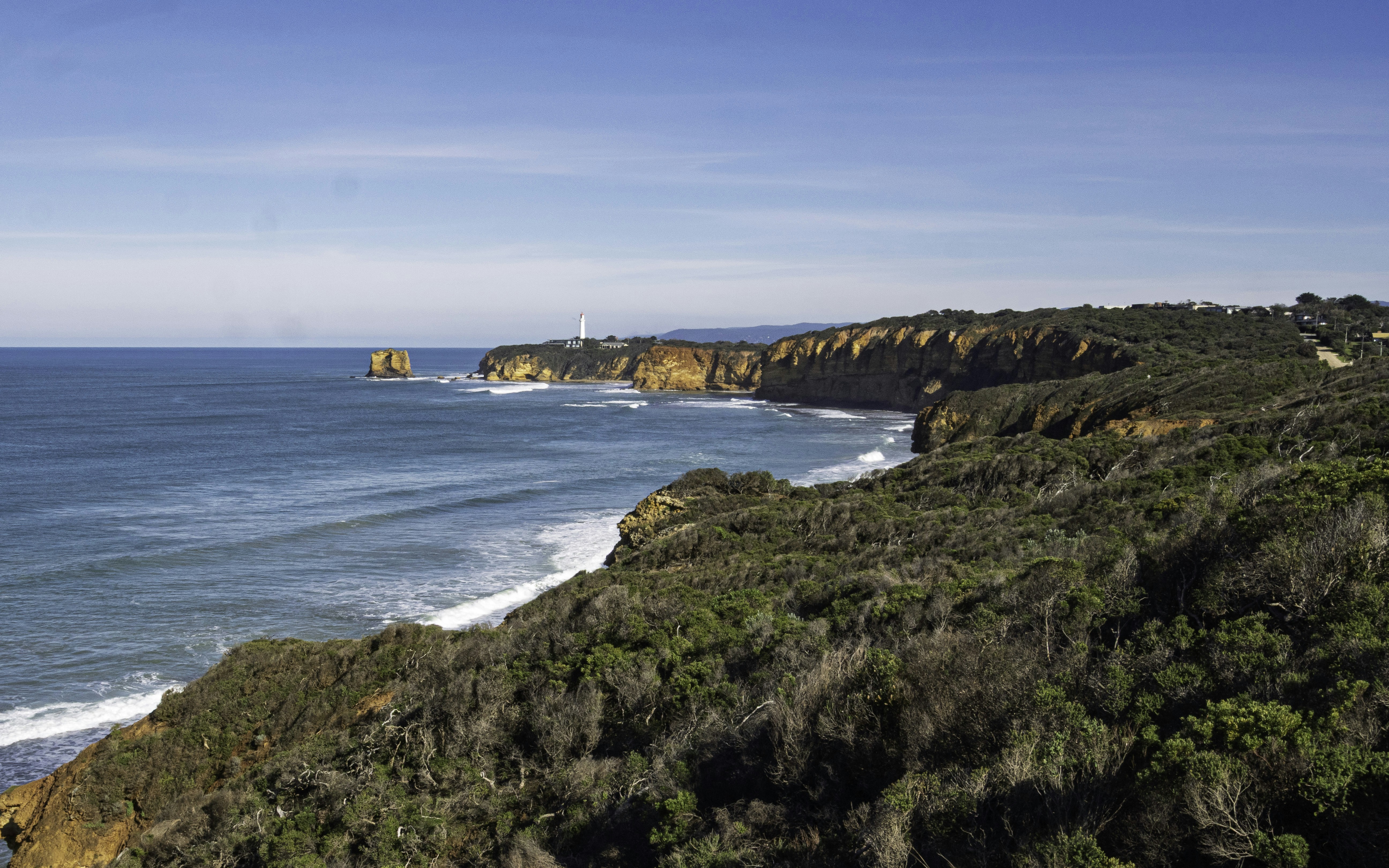 A rugged coastline with orange cliffs and a distant lighthouse under a clear blue sky
