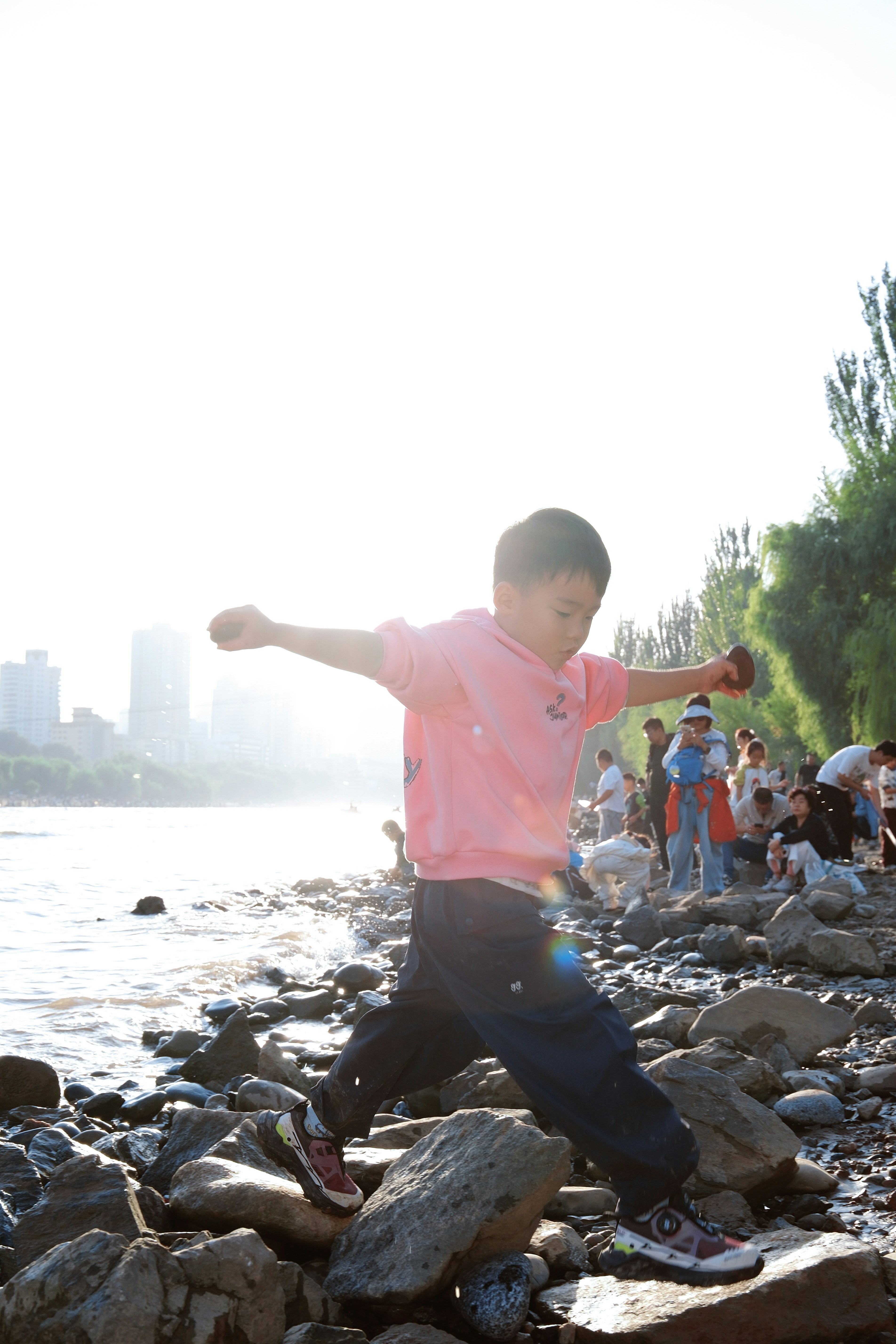 A child in a pink hoodie balancing on rocks along a riverbank in bright light