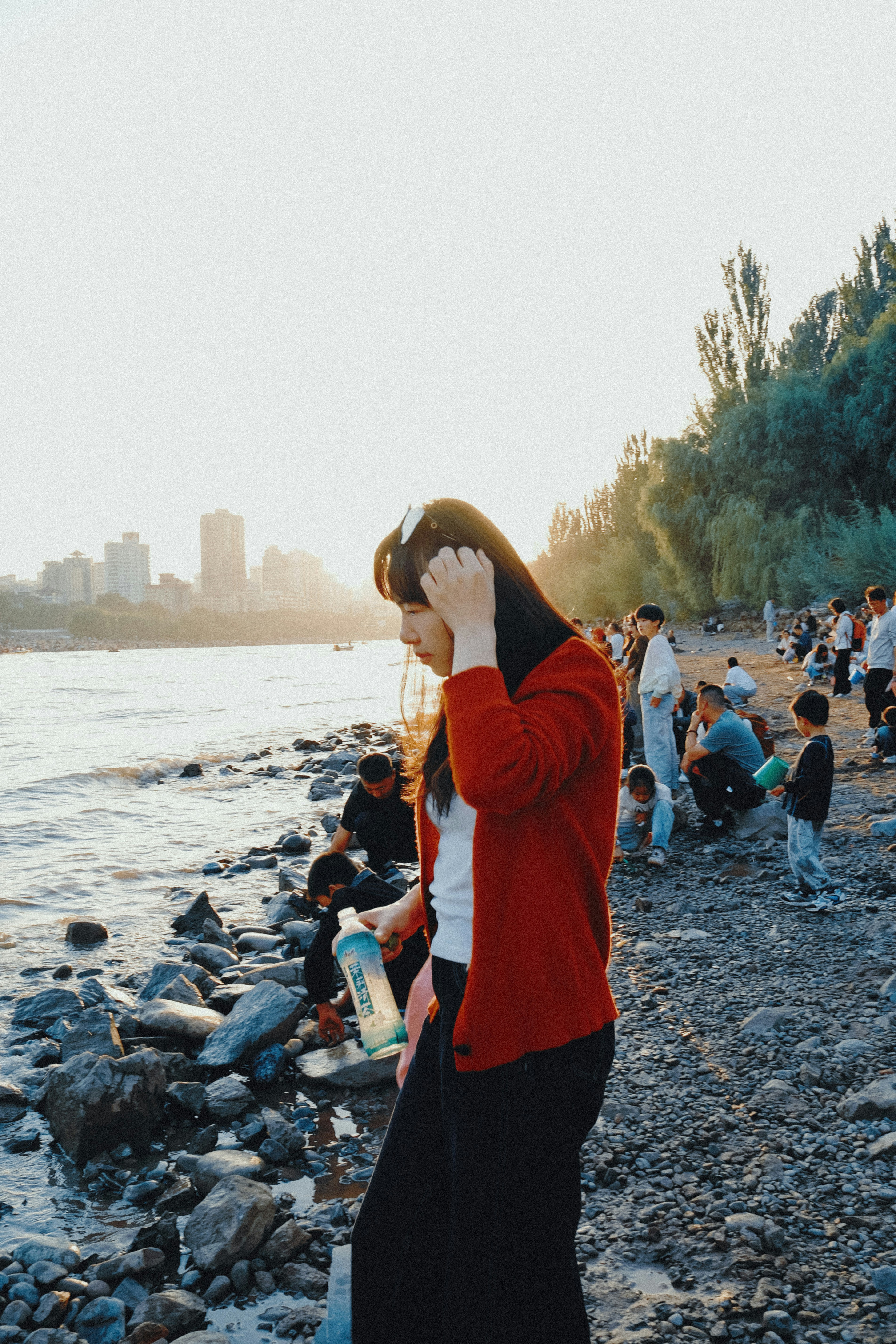 A woman in a red cardigan holding a water bottle on a rocky riverbank at golden hour