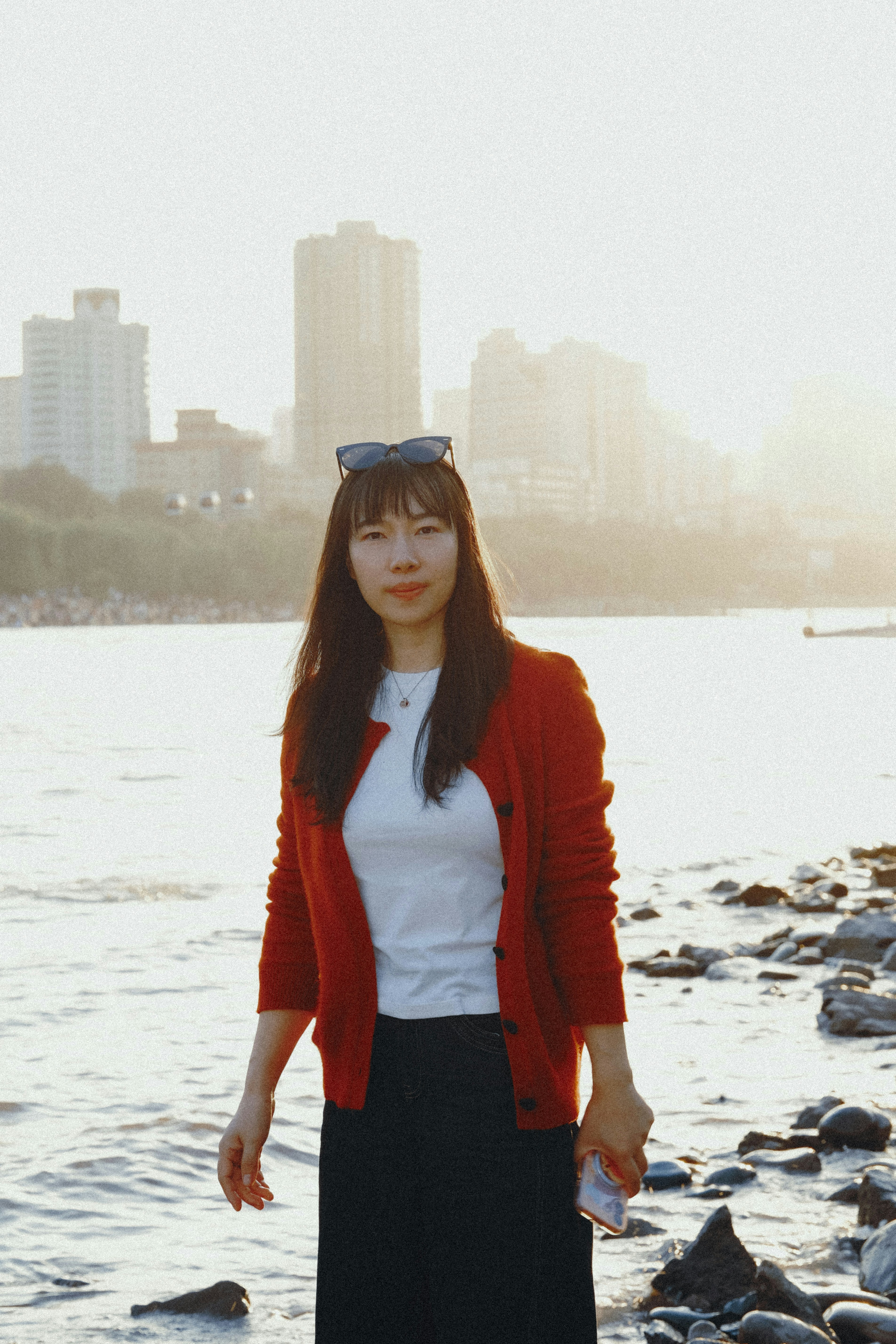A woman in a red cardigan standing by a river with city buildings in the background