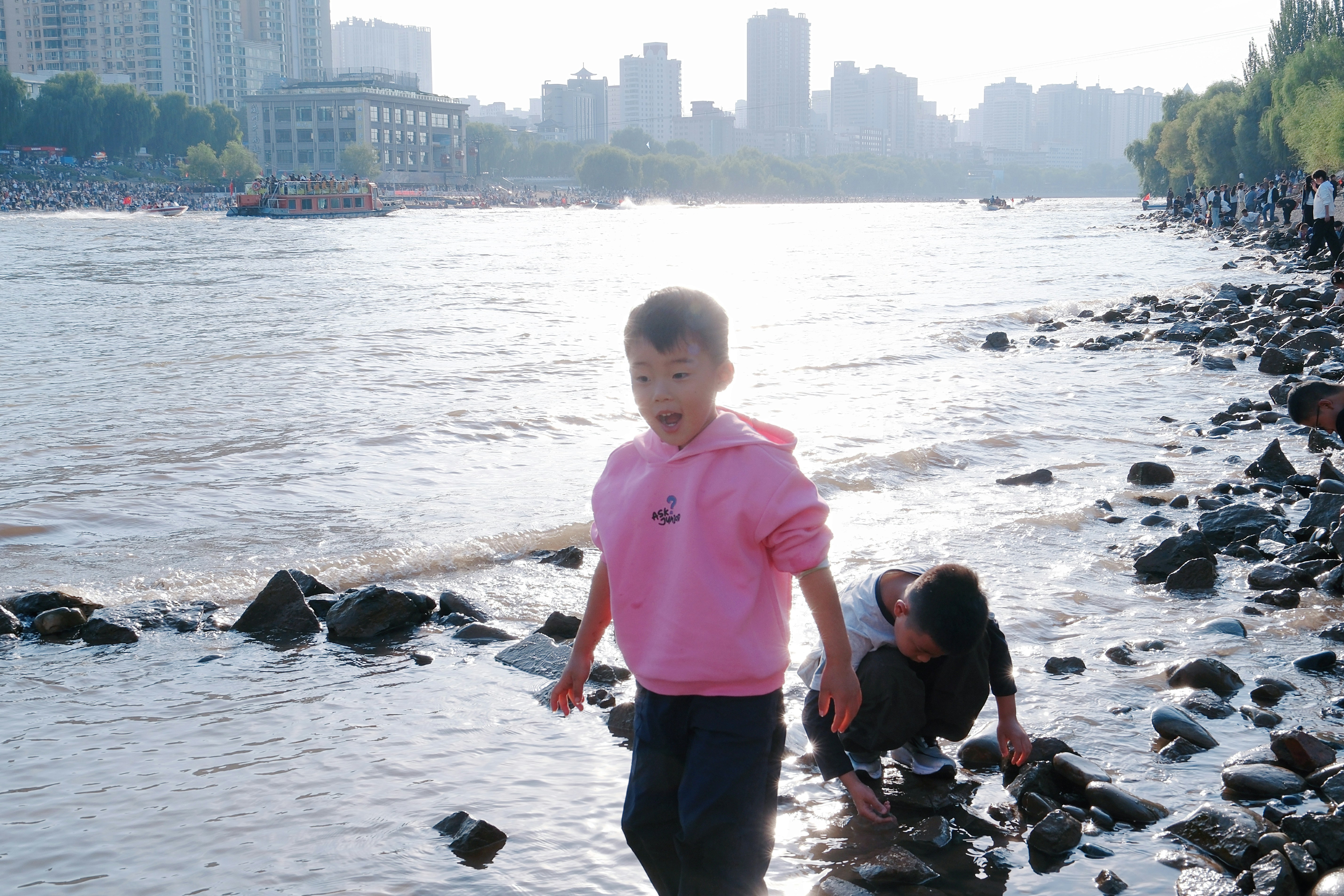 Two children playing on a rocky riverbank with a city skyline in the background