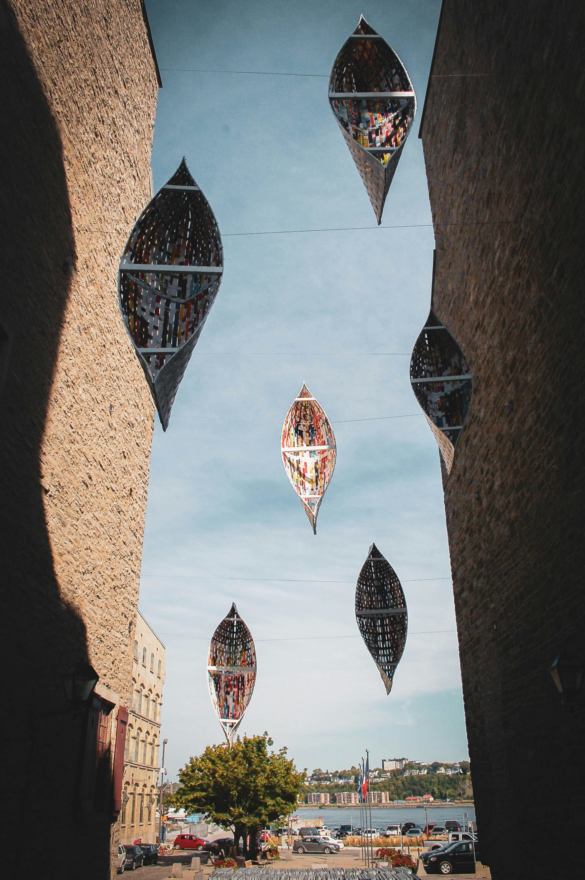 Suspended boat-shaped art installations hanging between two tall brick buildings above a street