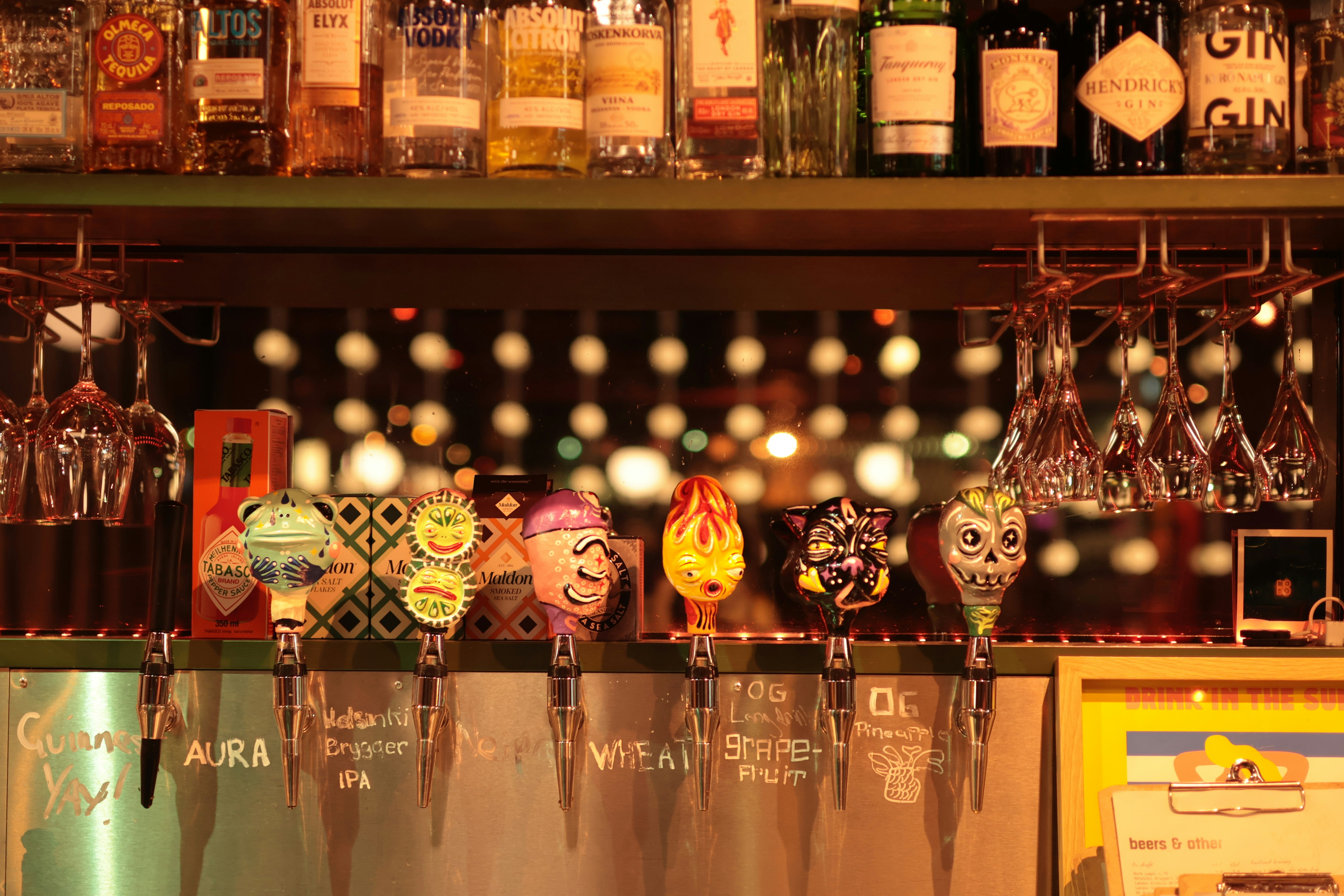 A row of colorful decorative beer tap handles on a metal bar counter with hanging glasses