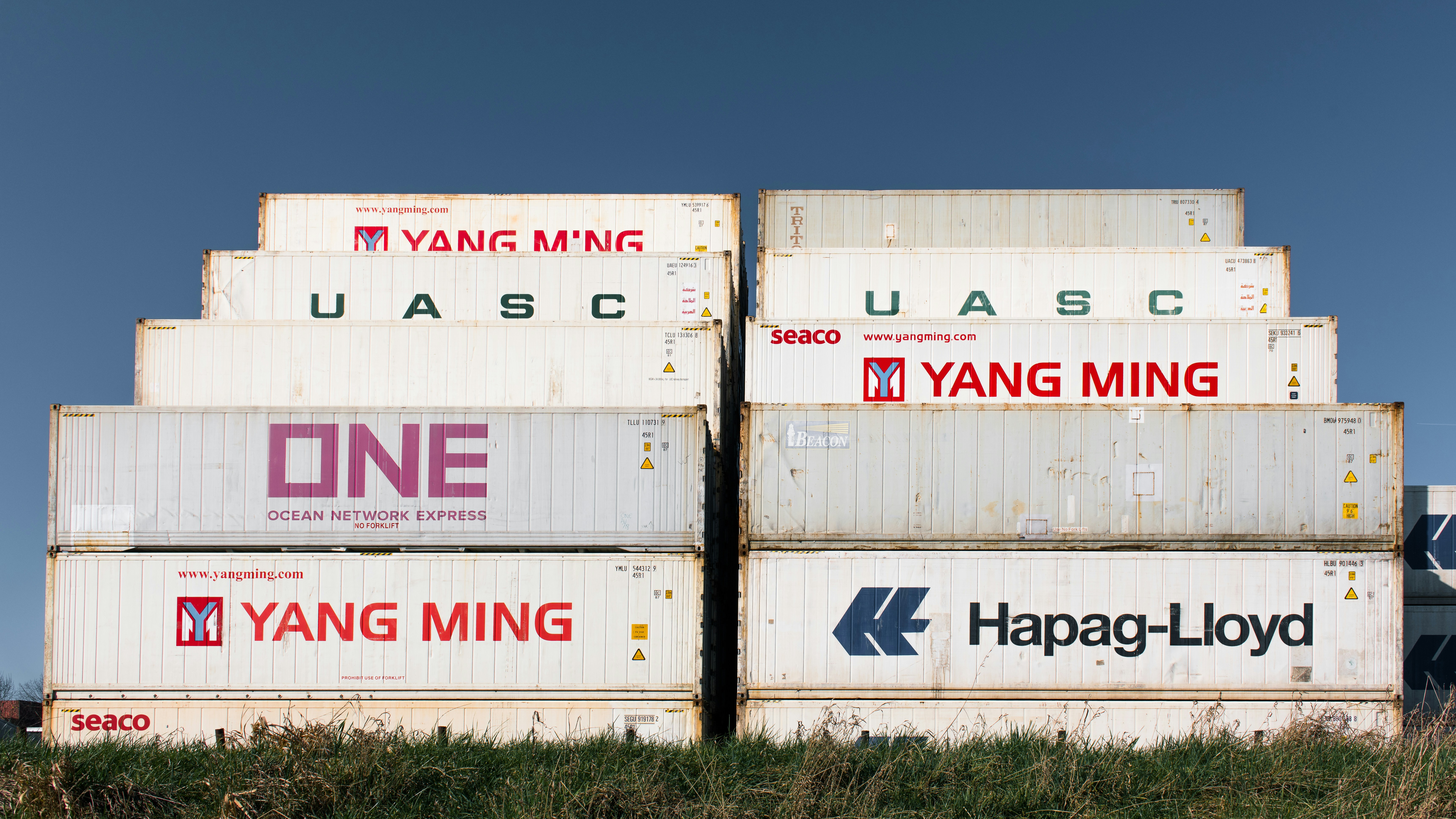 Stacked white shipping containers with various company logos against a clear blue sky