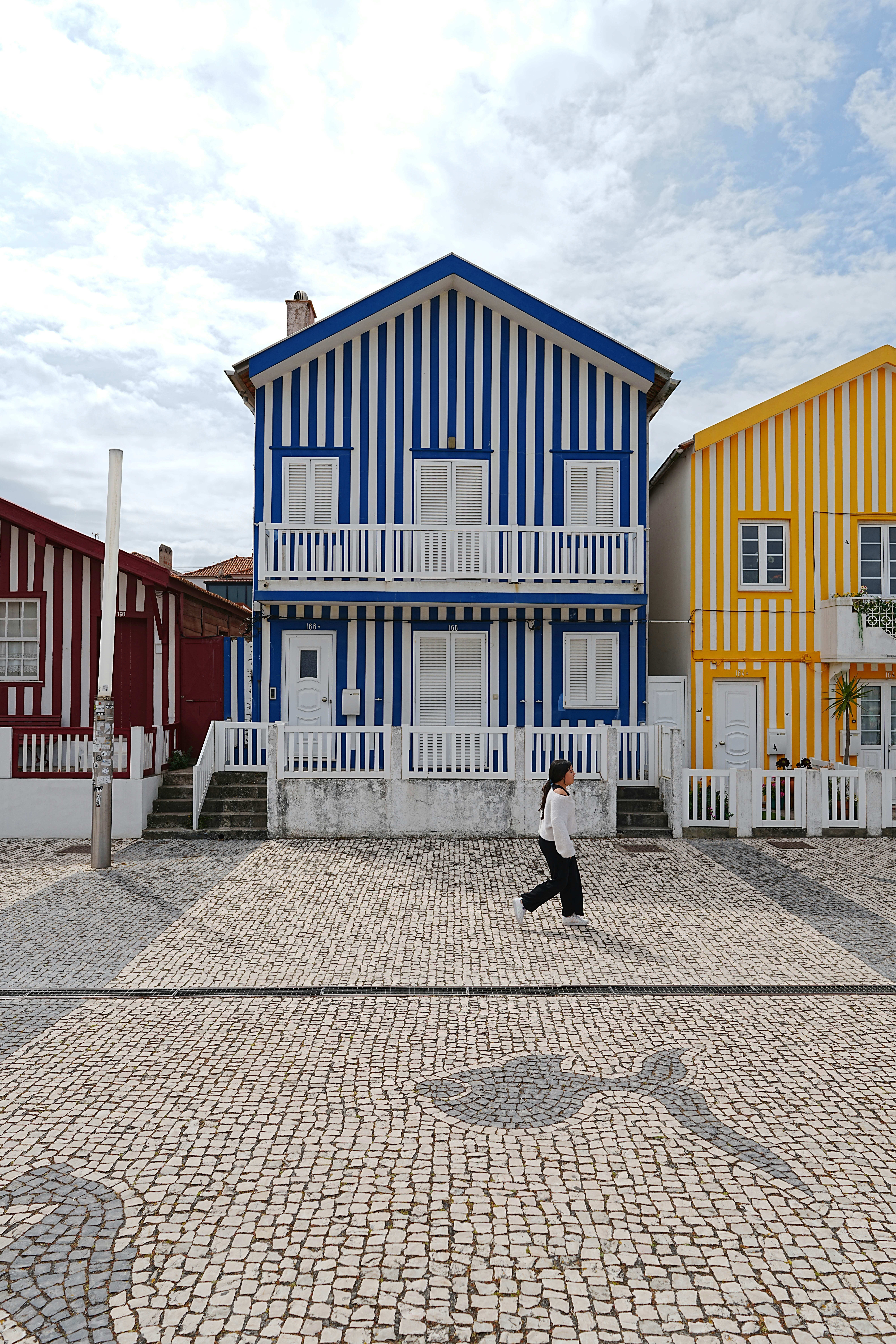 A person walking past blue and white striped houses on a cobblestone street in Costa Nova