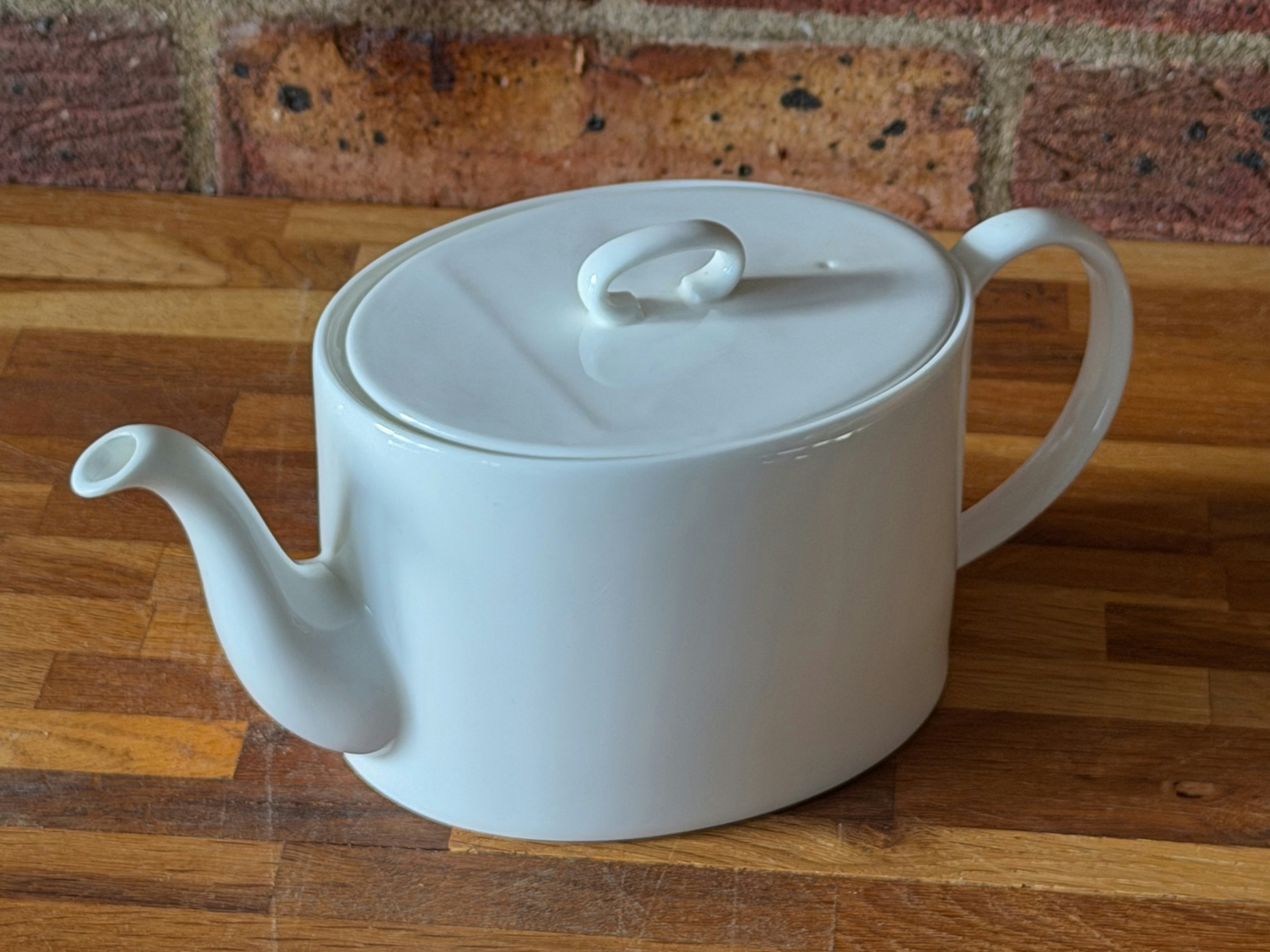A white ceramic teapot on a wooden surface against a brick wall