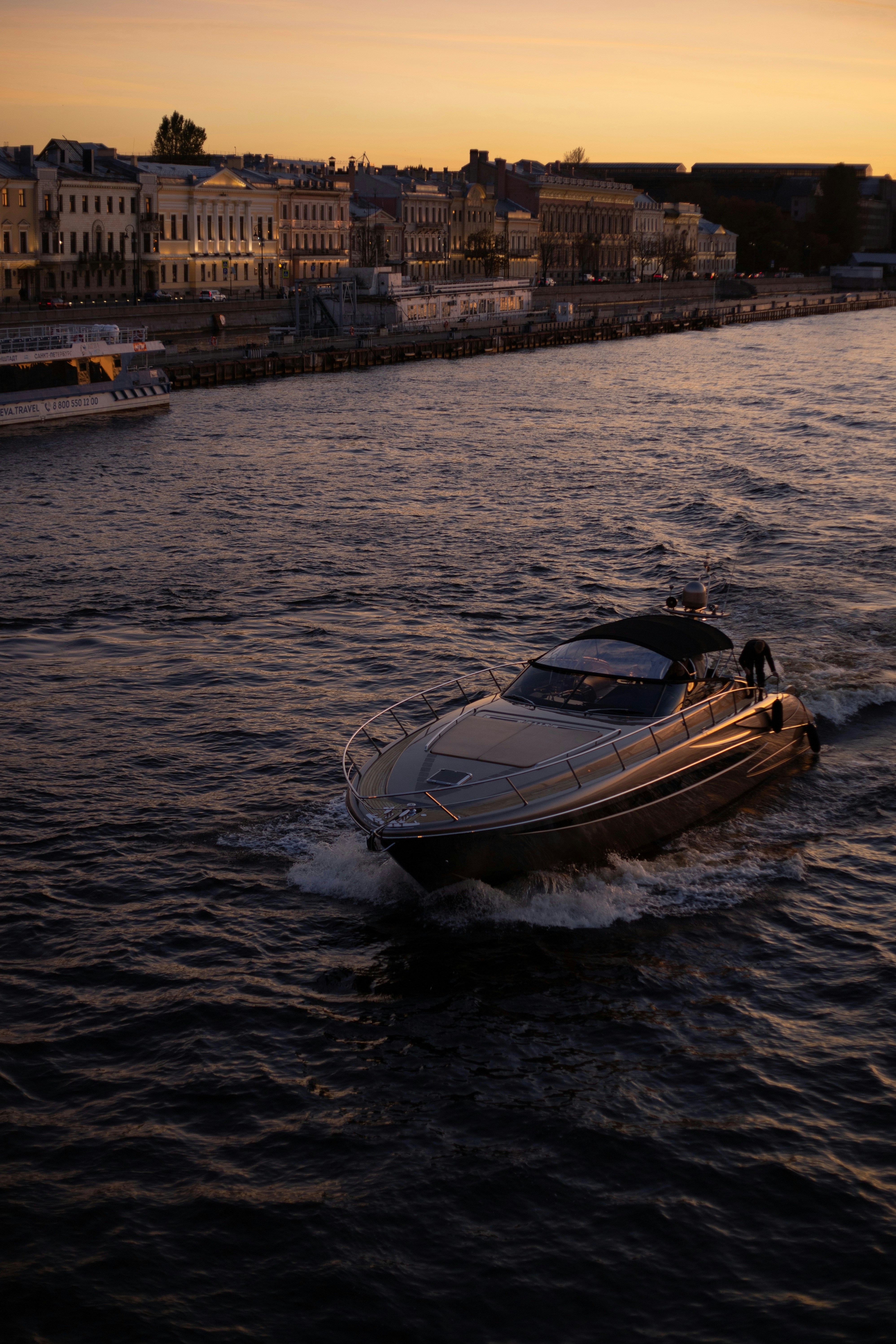 A motorboat cruising on a river past a city embankment during the golden hour