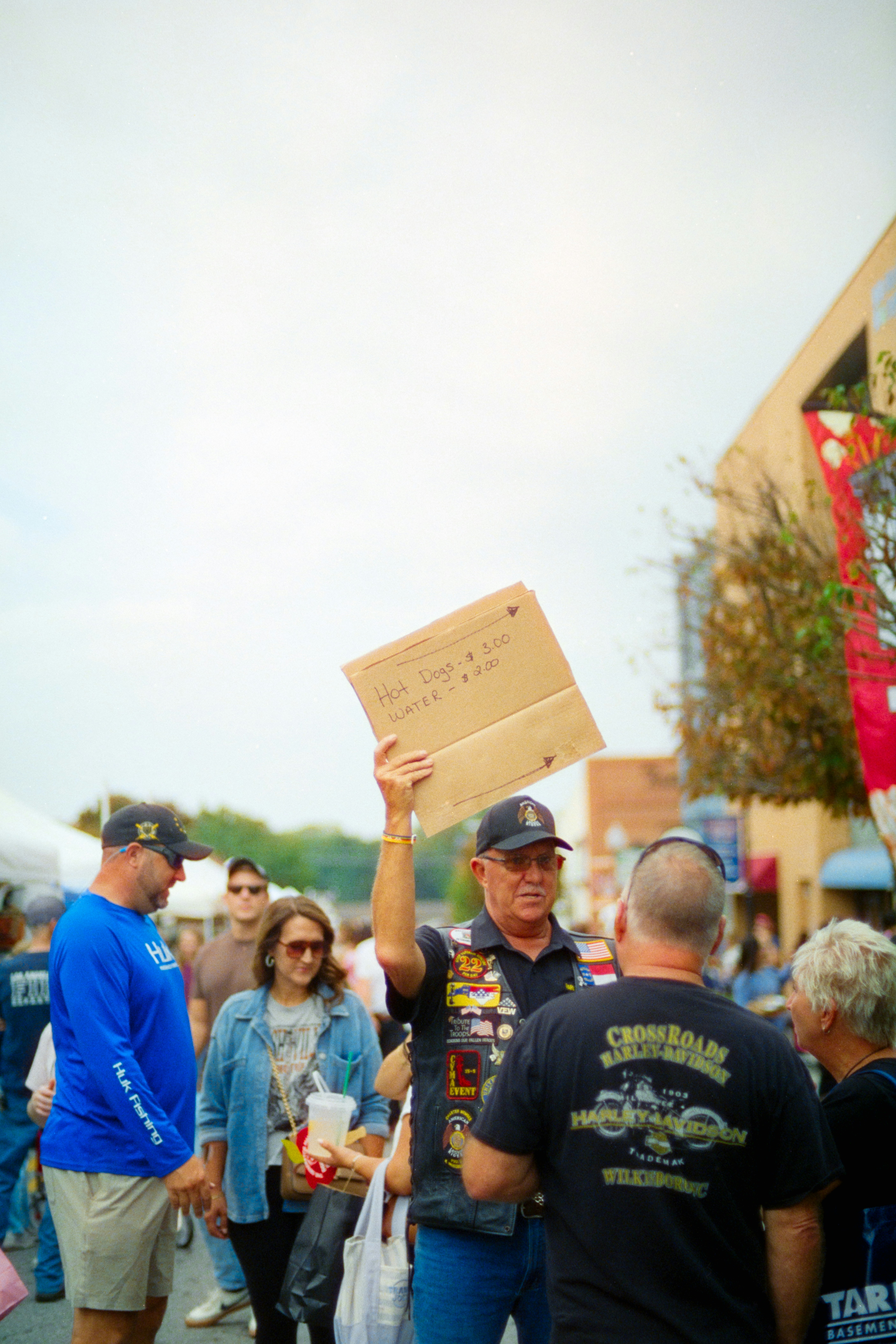A man in a vest holding a cardboard sign advertising hot dogs and water at an outdoor event