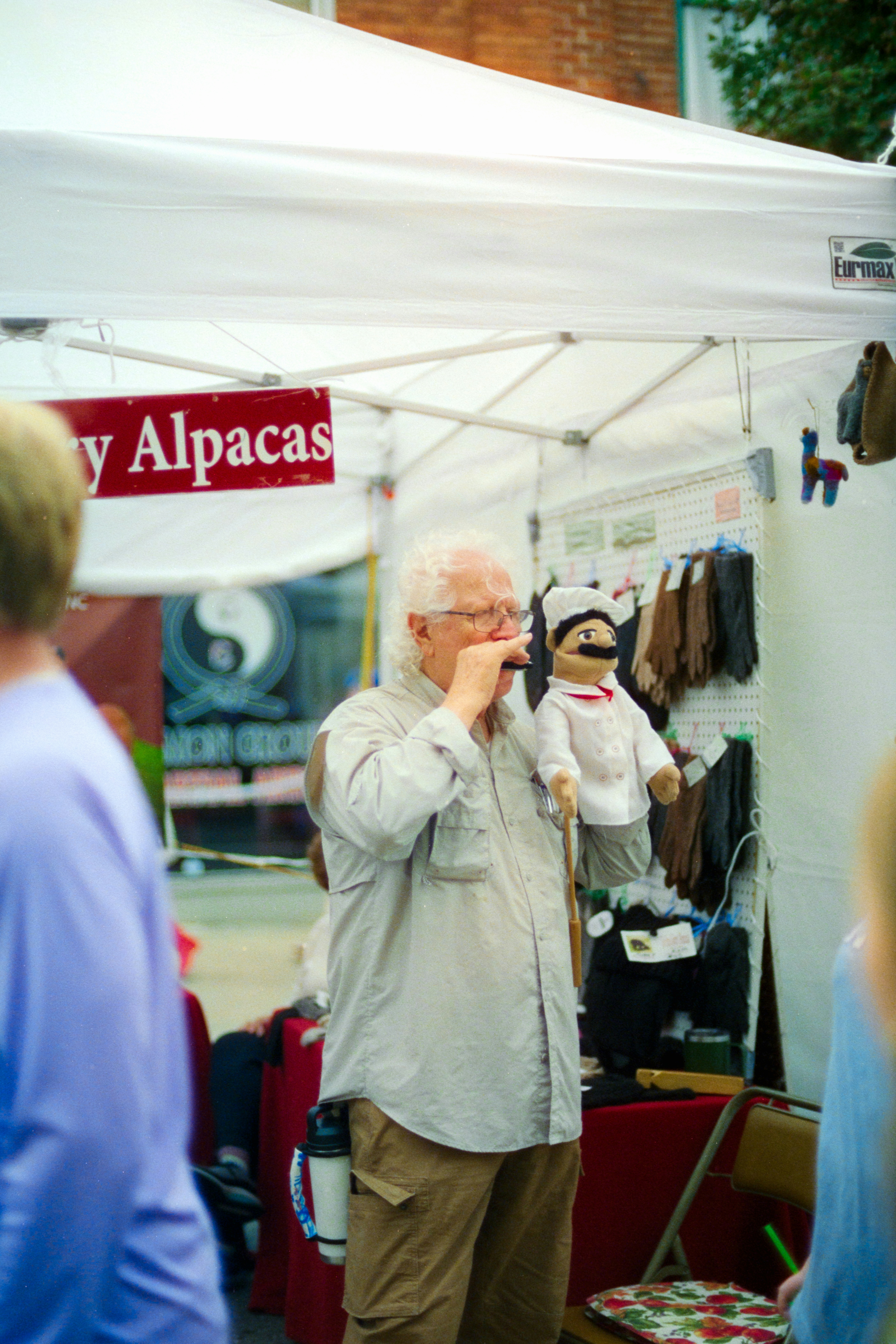 An adult man playing a harmonica while holding a chef puppet under a white market tent