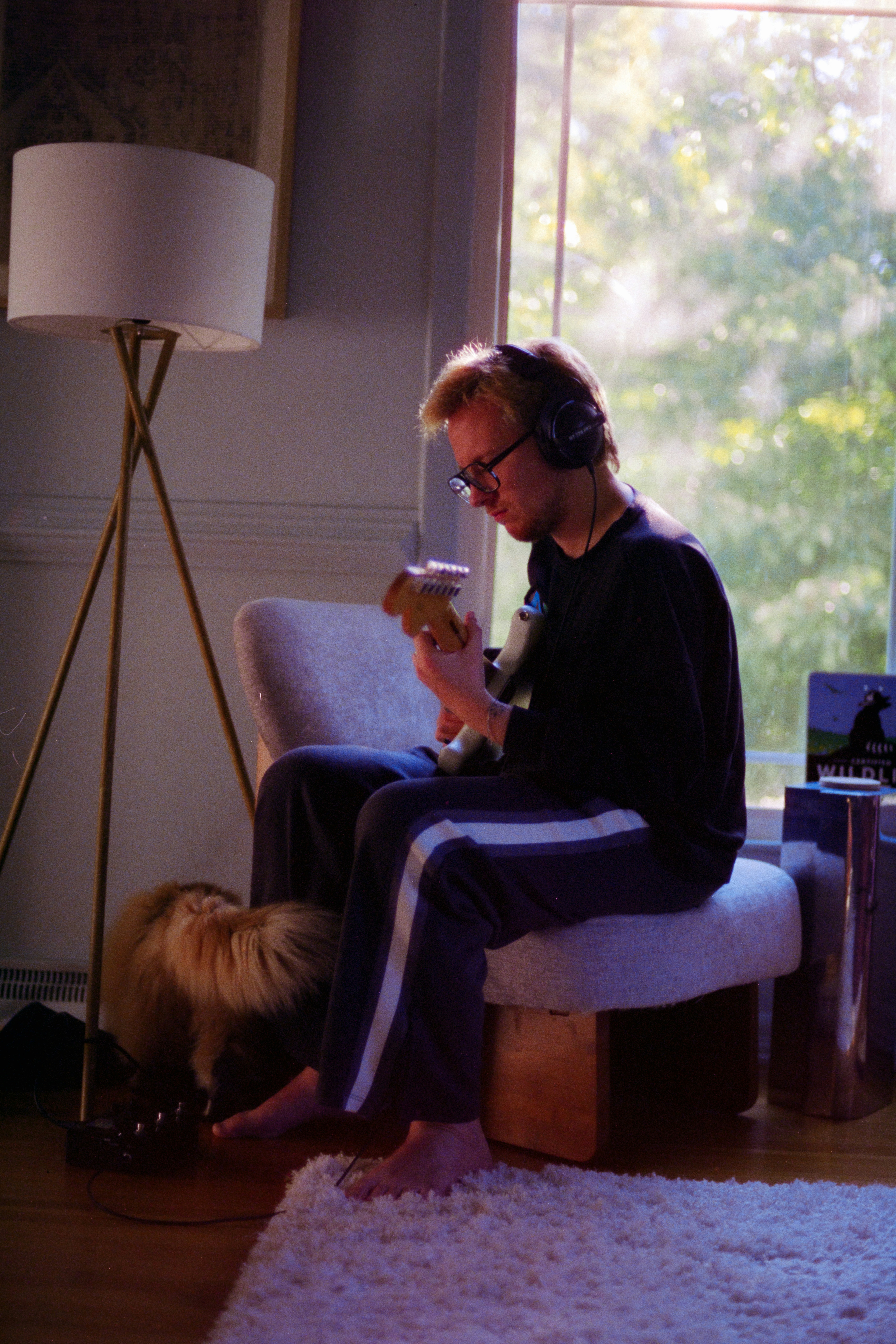 A person with headphones playing an electric guitar in a sunlit room with a floor lamp