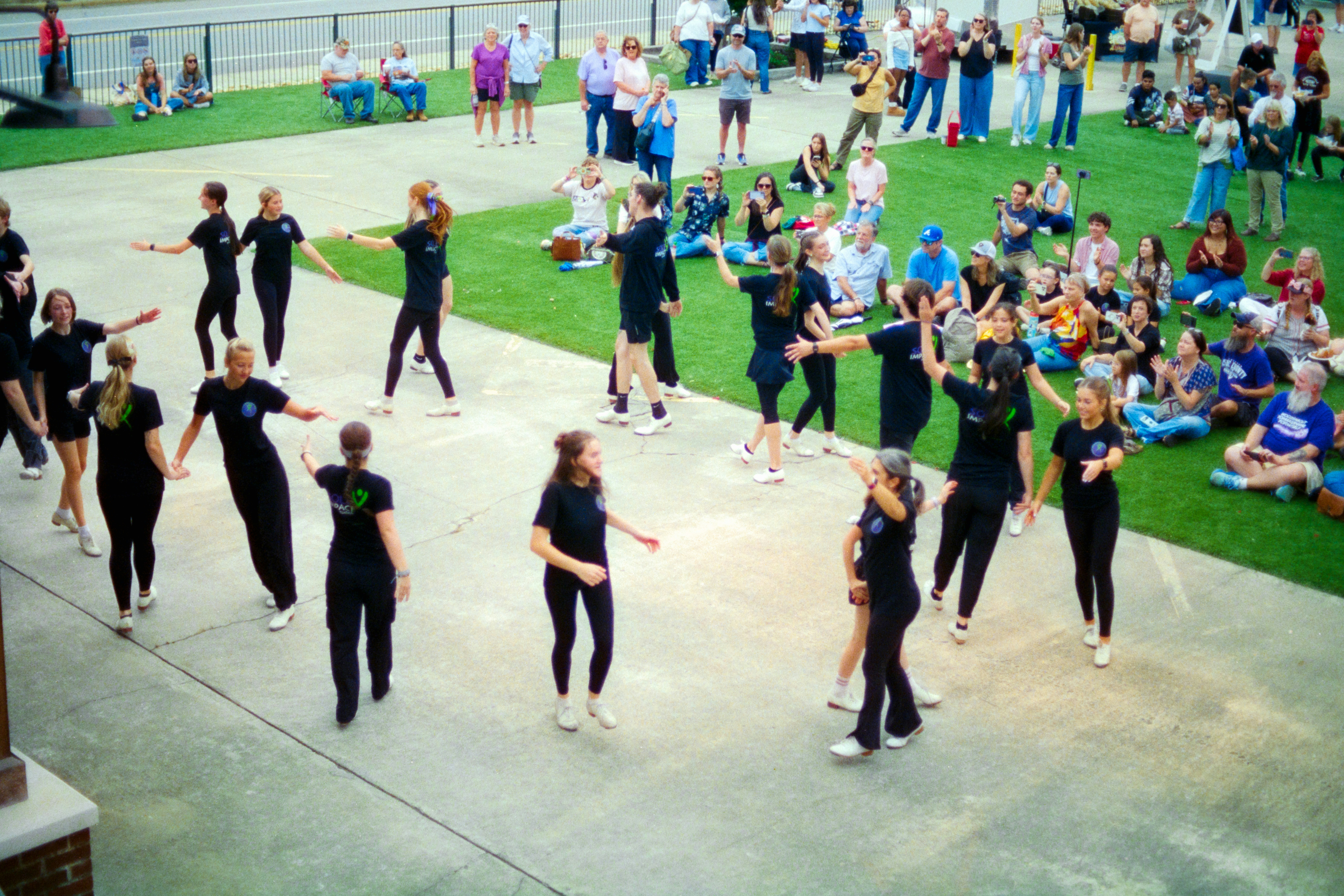 A group of dancers in black performing on a concrete plaza before an audience