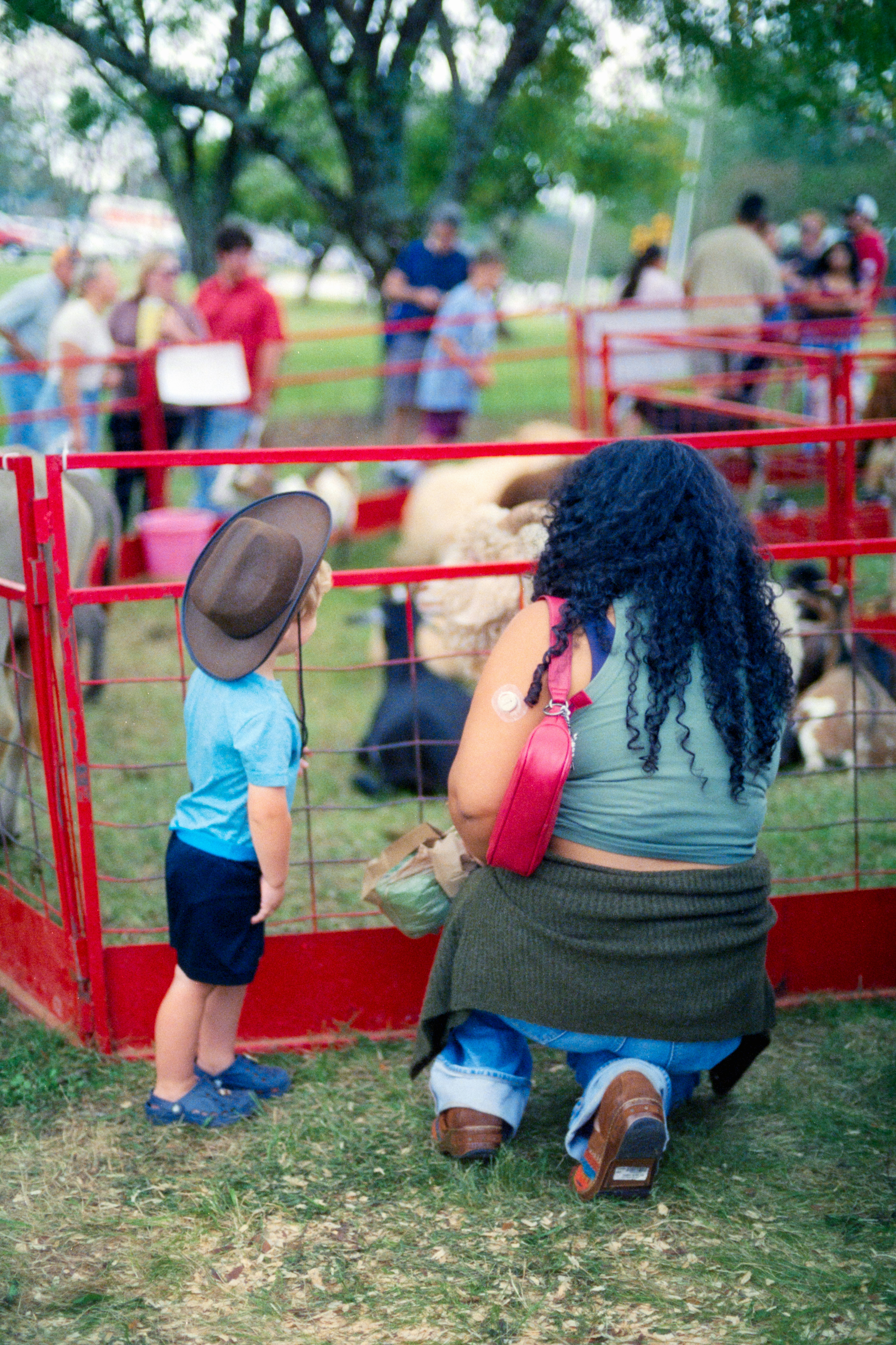 A child in a cowboy hat and an adult kneeling by a red fence at a petting zoo