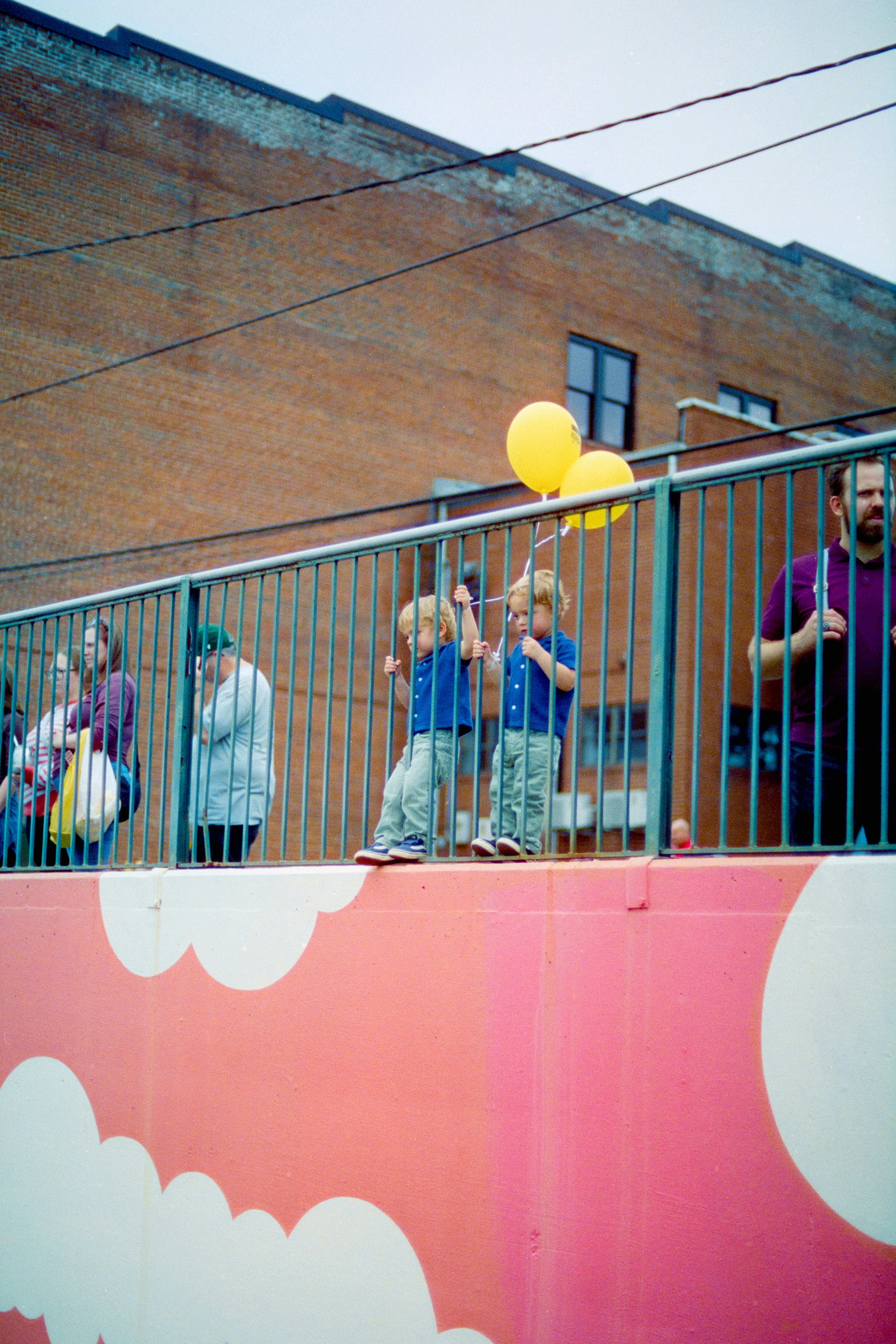 Two children holding yellow balloons while standing behind a metal railing on a pink wall