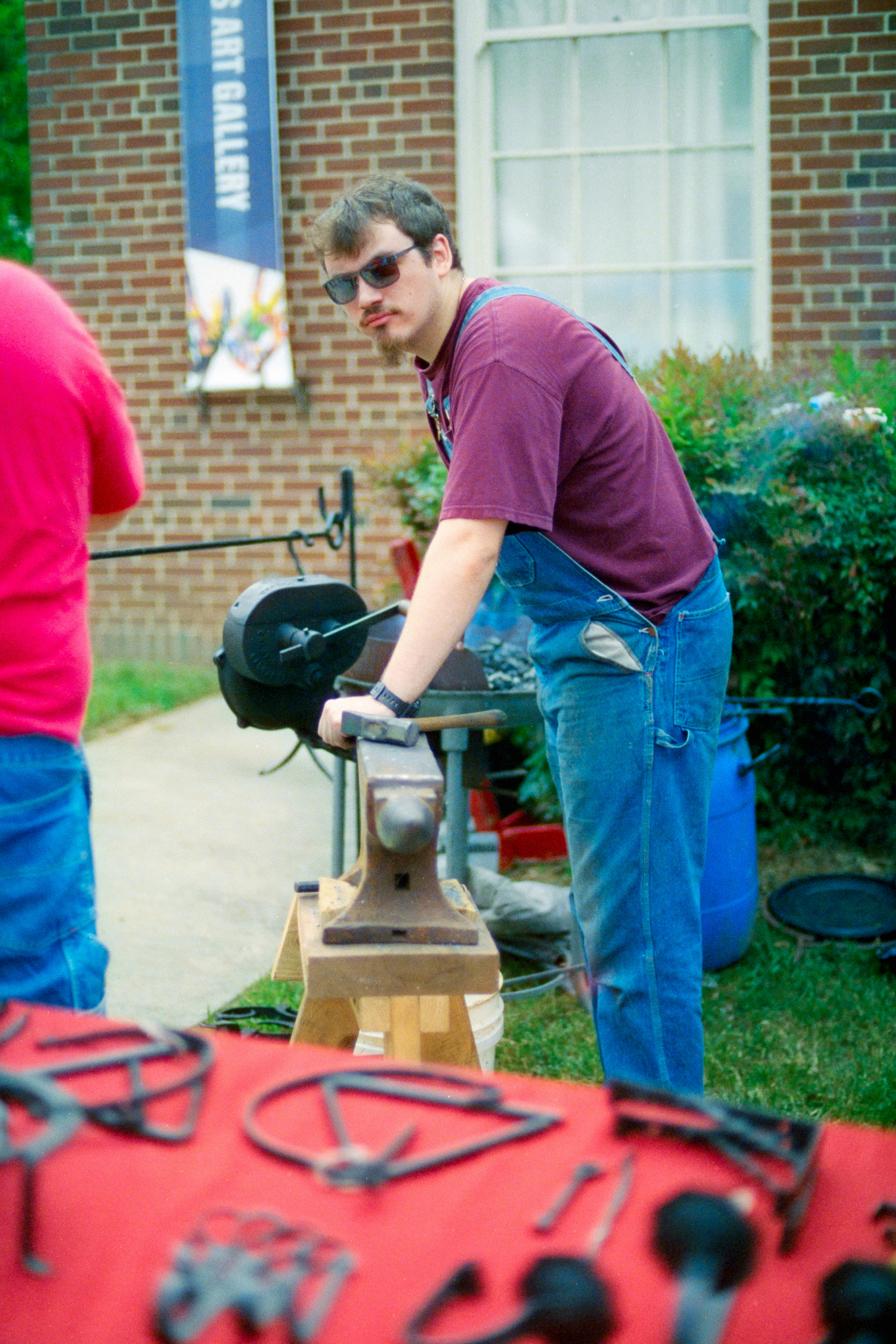 A man in denim overalls and a maroon t-shirt working at an anvil outdoors